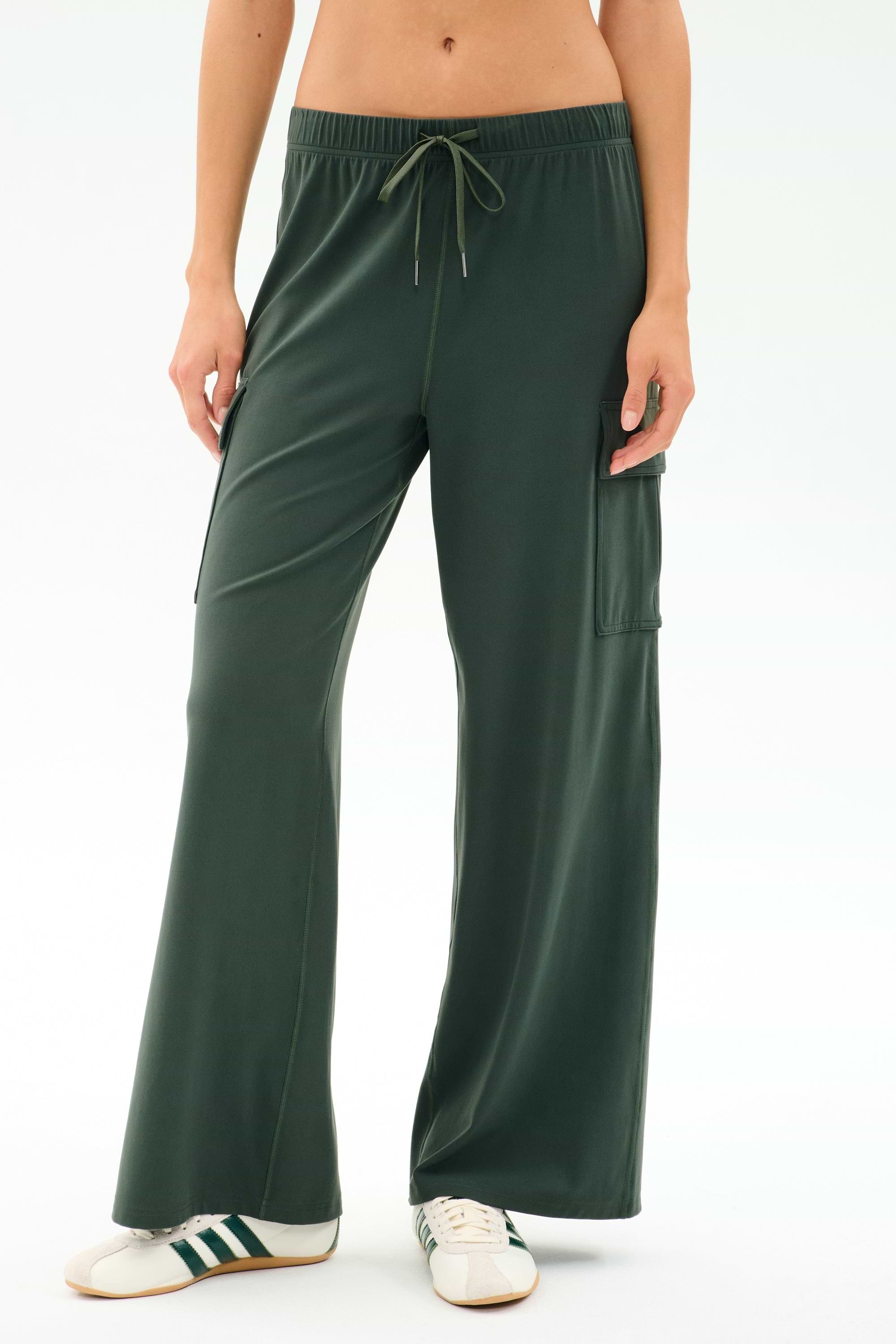 A person wearing SPLITS59 Alex Airweight Cargo Pant - Military, dark green wide-leg spandex blend pants with a side pocket, paired with white sneakers featuring green stripes, shown from waist to feet.