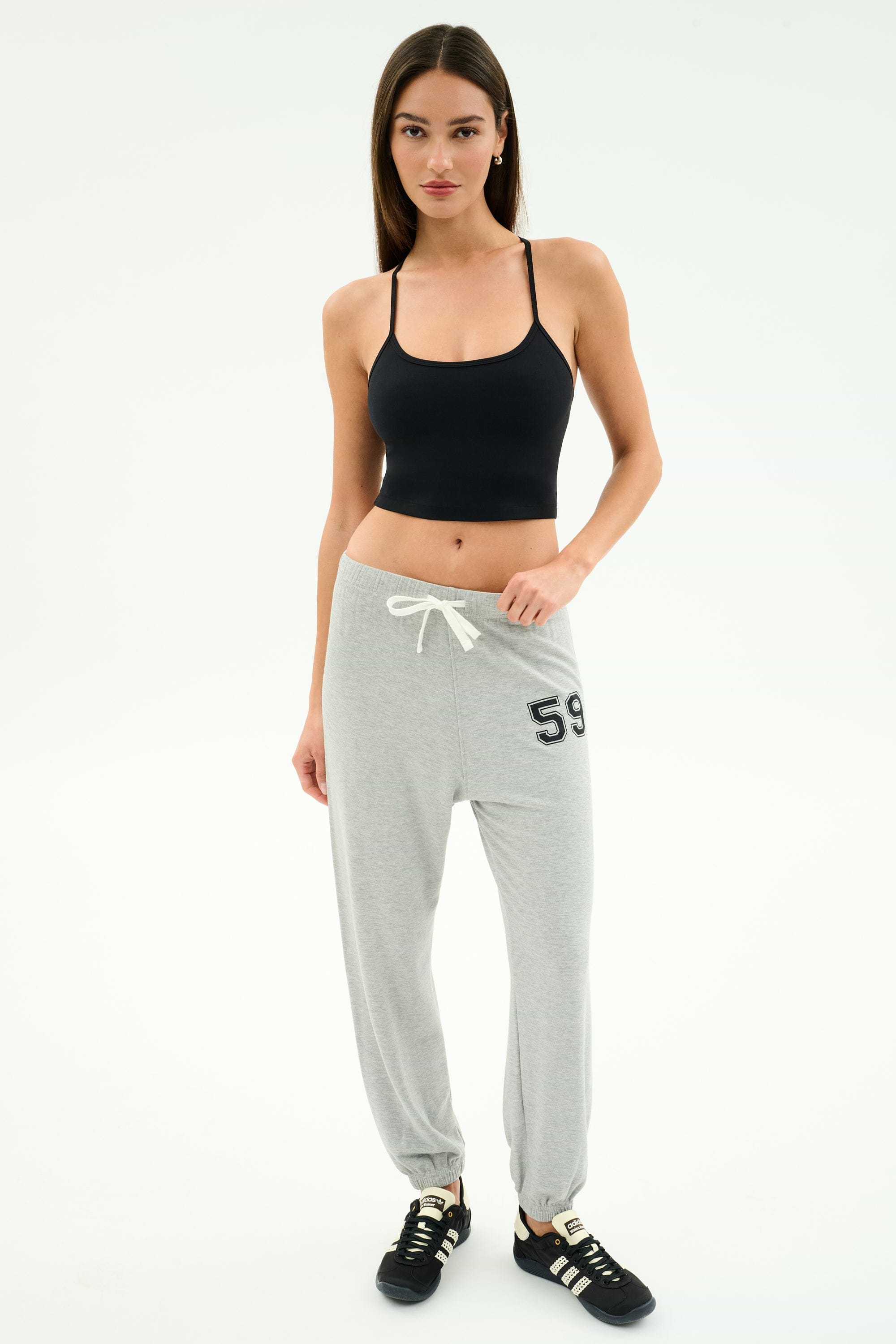 A woman stands against a plain background wearing a black tank top, SPLITS59 Andie Oversized Fleece Varsity 59 Sweatpant in Heather Grey with "59" on the hip, and black sneakers.