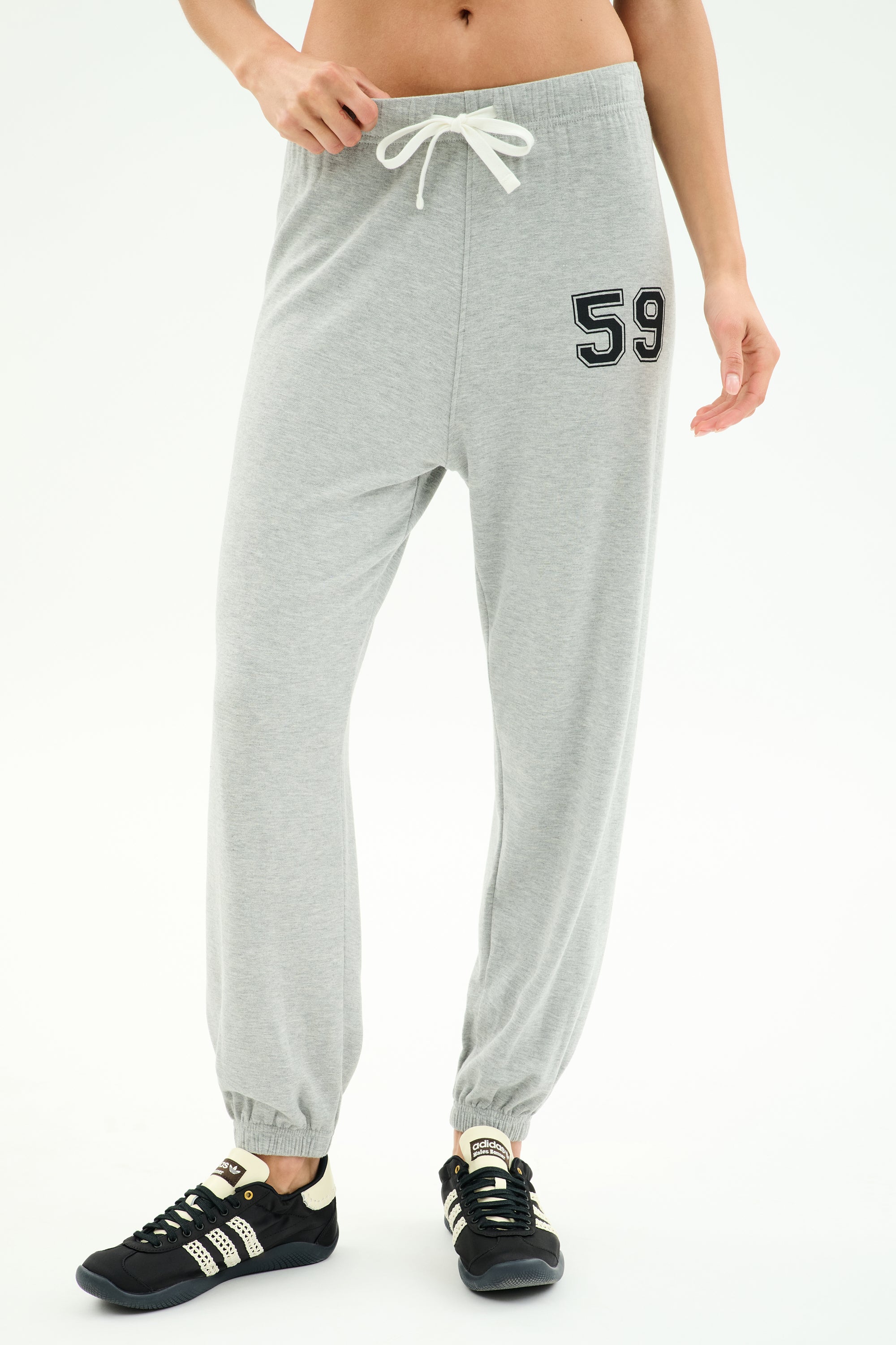 A person wears SPLITS59 Andie Oversized Fleece Varsity 59 Sweatpant in Heather Grey/59, featuring a drawstring waist and "59" patch on the left thigh, paired with black sneakers. Only the lower torso and legs are visible. Made in Los Angeles.