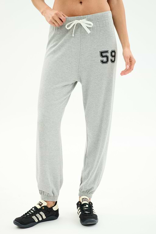 A person wears SPLITS59 Andie Oversized Fleece Varsity 59 Sweatpant in Heather Grey/59, featuring a drawstring waist and "59" patch on the left thigh, paired with black sneakers. Only the lower torso and legs are visible. Made in Los Angeles.