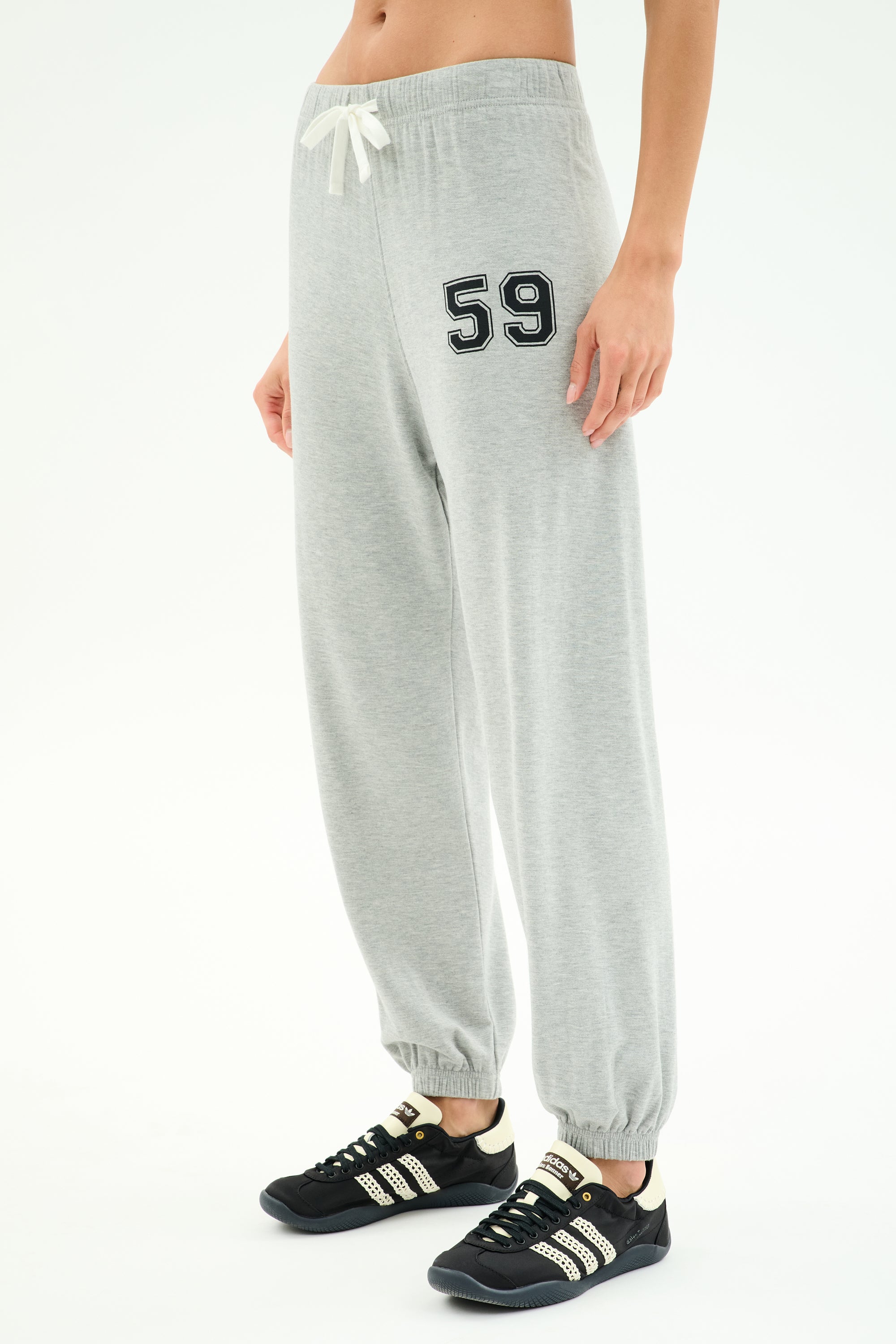 A person wearing SPLITS59 Andie Oversized Fleece Varsity 59 Sweatpant in Heather Grey/59, featuring "59" on the left thigh, white drawstring, and paired with black athletic shoes with white stripes.
