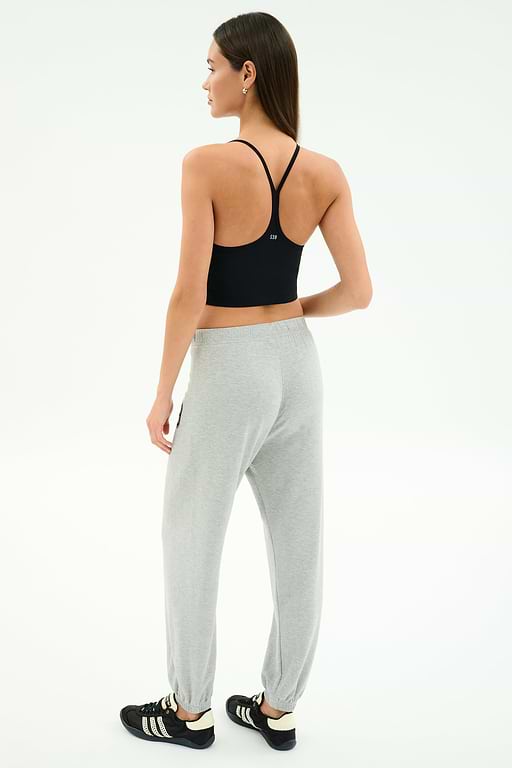 A woman faces away wearing a black sports bra, SPLITS59 Andie Oversized Fleece Varsity 59 Sweatpant in Heather Grey/59, and black athletic shoes with white stripes against a plain white background.
