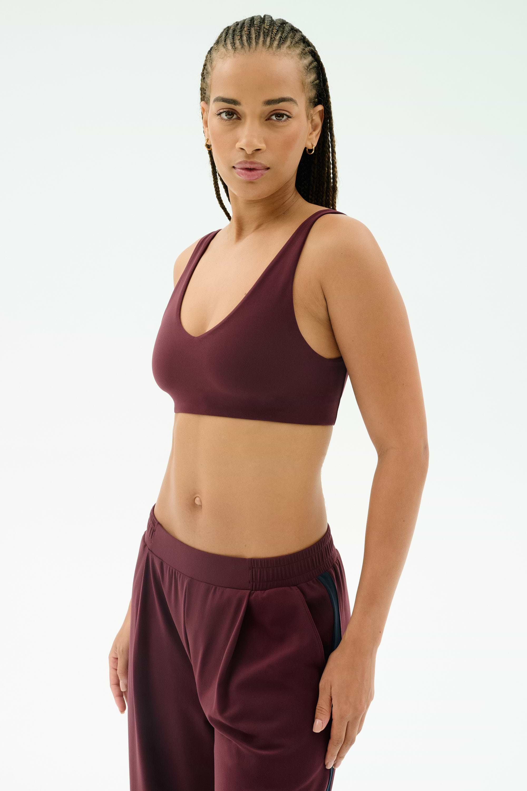 A woman with braided hair wears the SPLITS59 Airweight Scoop Back Bra in Dark Cherry, paired with matching track pants, made from nylon spandex, standing against a plain light background.