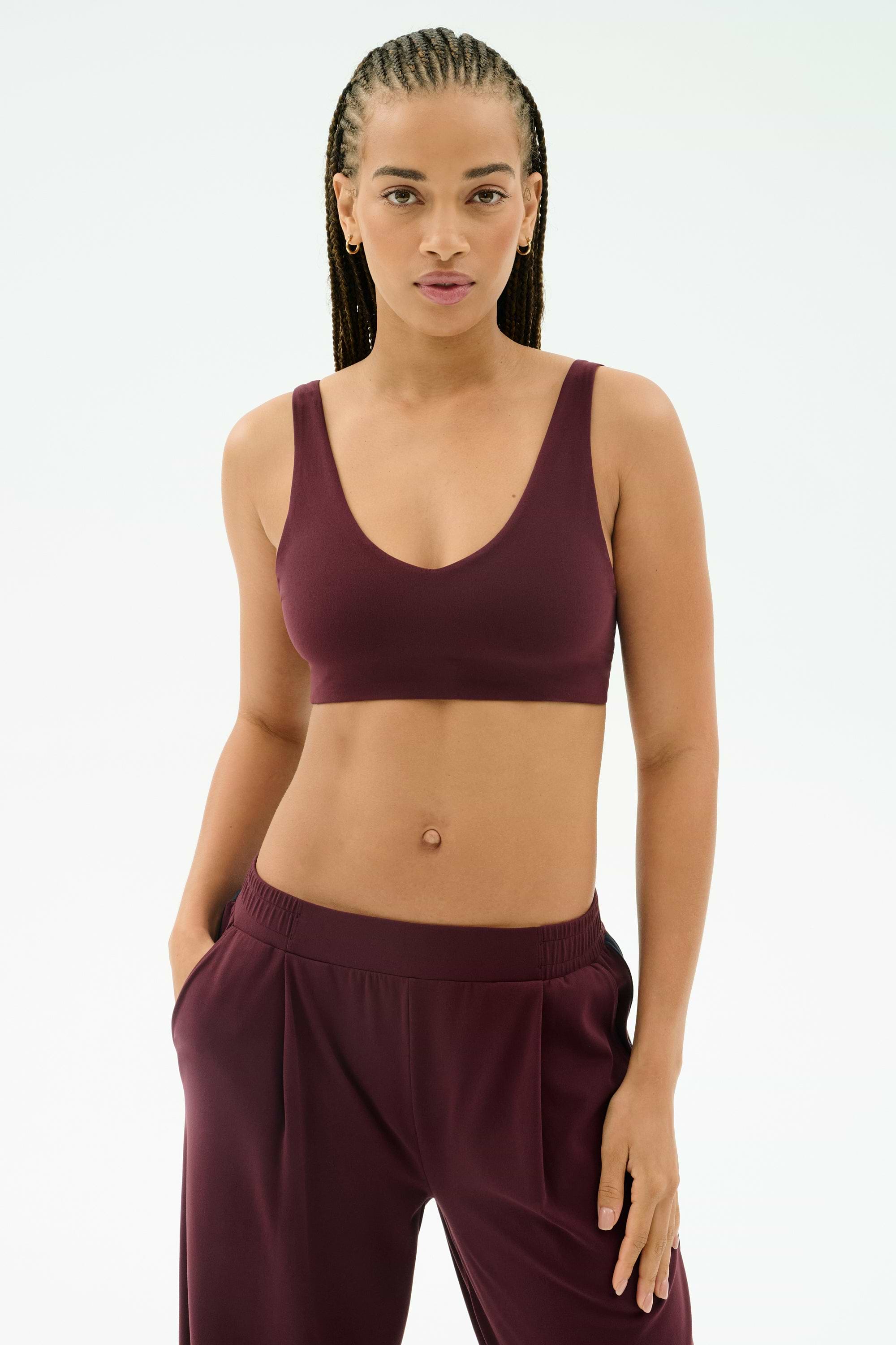 A woman wears the SPLITS59 Airweight Scoop Back Bra in Dark Cherry, made from soft nylon spandex, paired with matching pants and stands against a plain light background with one hand in her pocket.