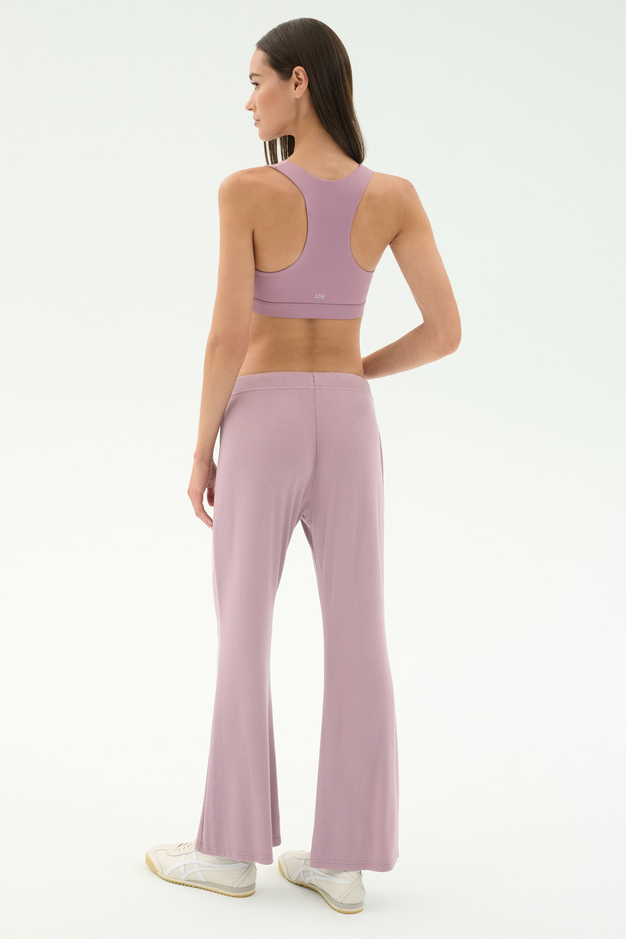 A woman faces away, wearing the SPLITS59 Blaire Rib Pant in blush with a matching lavender sports bra and white sneakers, set against a plain, light background. The outfit is crafted from soft modal fabric with added spandex for comfort.