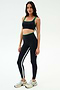 A woman stands against a plain background wearing the SPLITS59 Cait Rigor Bra in Black/Grass, crafted from high-performance Rigor fabric—perfect for high-impact workouts—paired with black leggings and white sneakers.
