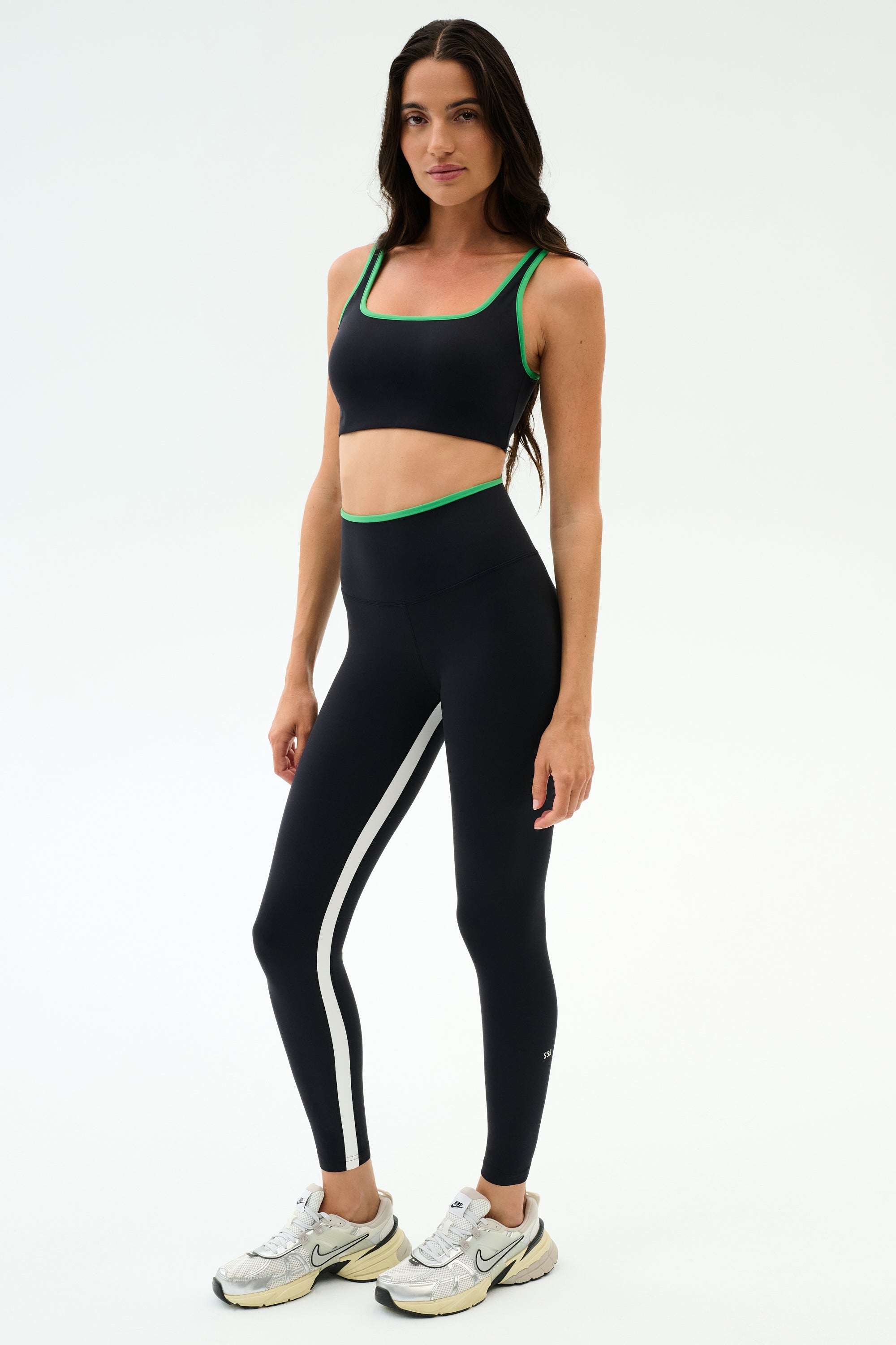 A woman stands against a plain background, wearing the SPLITS59 Easton Rigor High Waist 7/8 leggings in Black/Grass with green trim, paired with a black sports bra and white sneakers. The compressive fabric offers workout support.