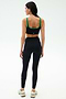 A woman faces away, wearing the SPLITS59 Cait Rigor Bra in Black/Grass, made from high-performance Rigor fabric, paired with high-waisted black leggings and white sneakers against a plain white background.