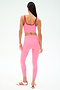 A woman faces away, wearing the SPLITS59 Cait Rigor Bralette in Strawberry/Pirate Red with matching pink leggings and white sneakers with red stripes, set against a plain background.