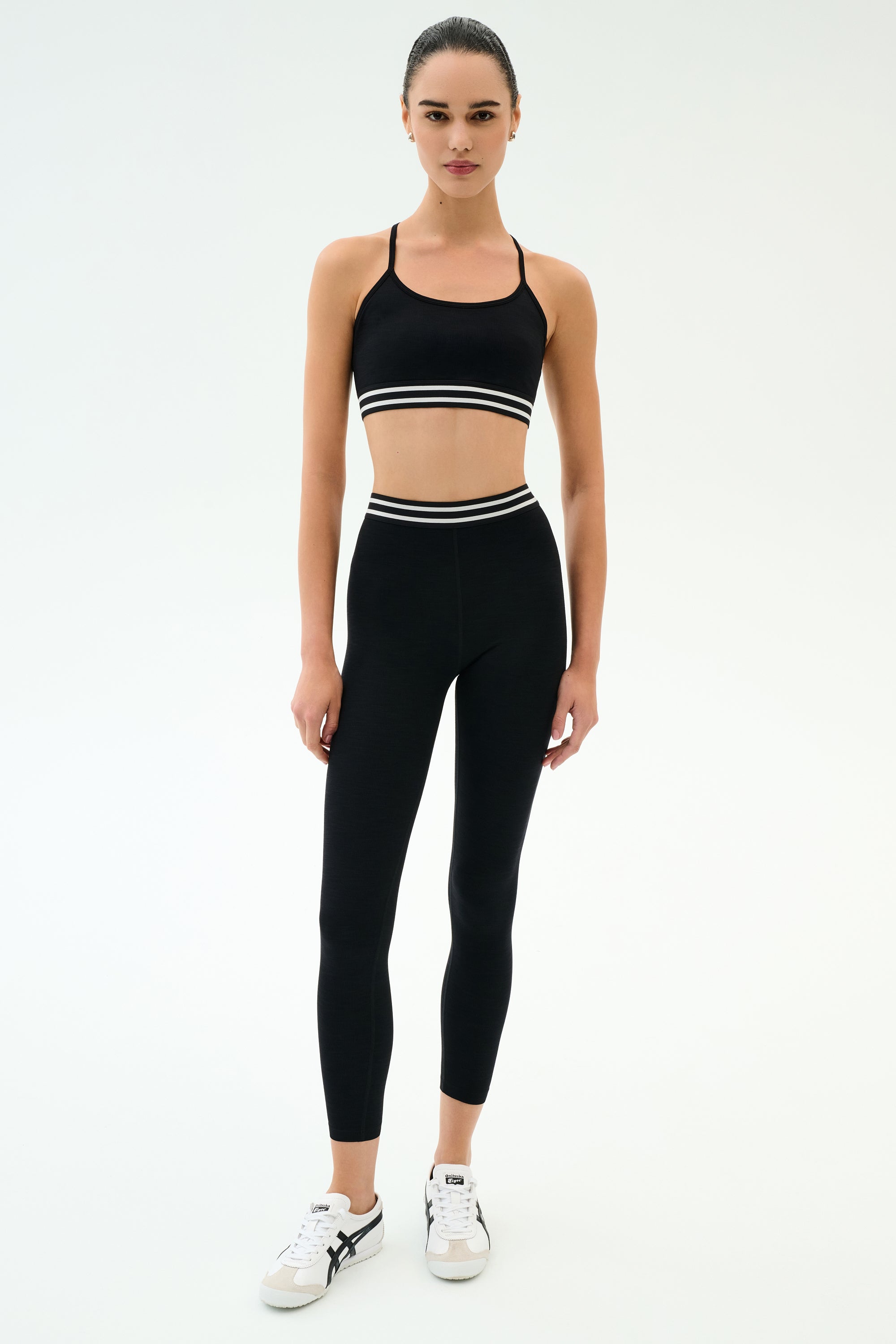 A person wearing the SPLITS59 Cali Active Rib Bra in black and high-waisted black leggings with white stripes, paired with white sneakers, stands against a plain white background.