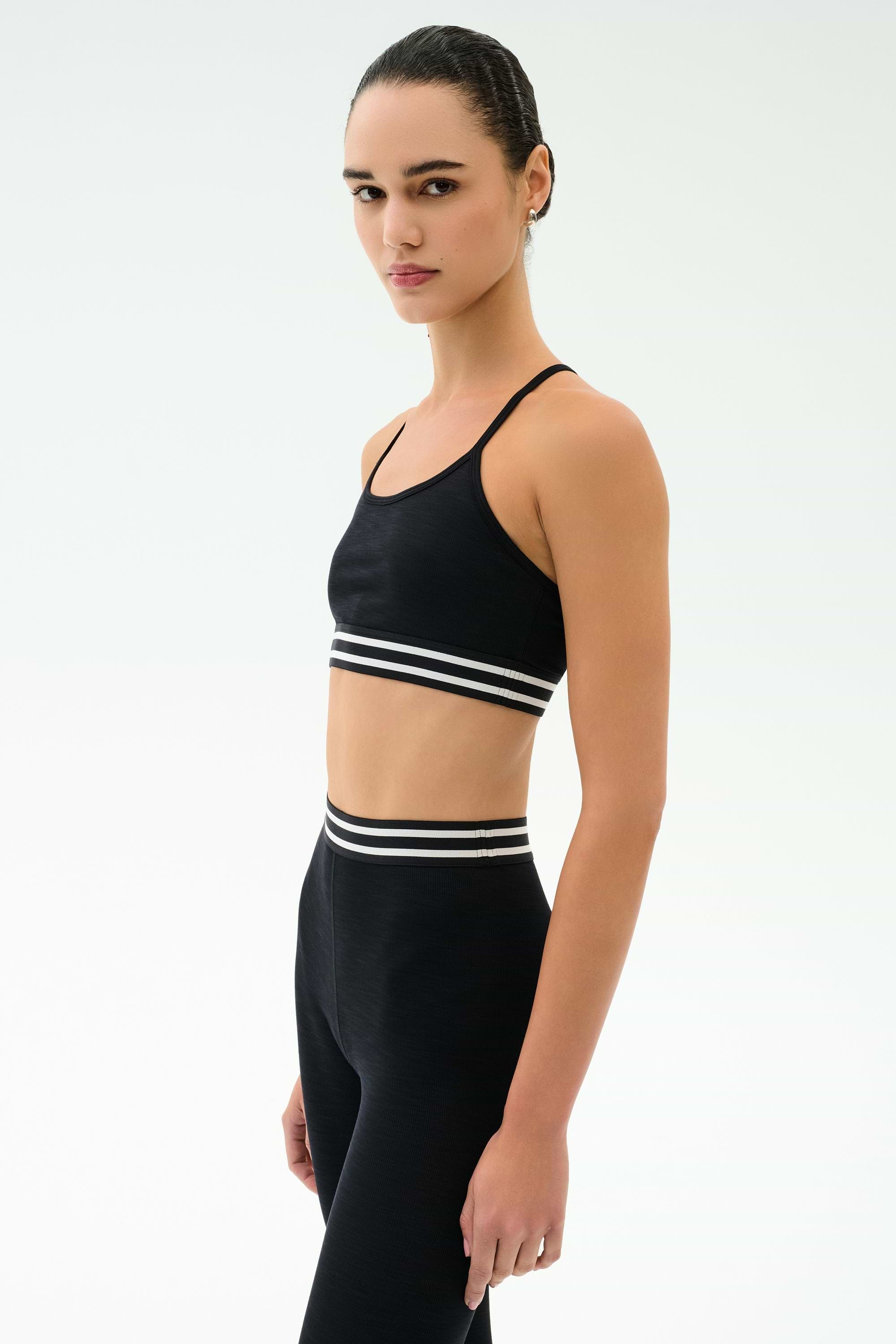 A woman stands sideways against a white background, wearing the SPLITS59 Cali Active Rib Bra in black and matching high-waisted leggings with white striped bands.