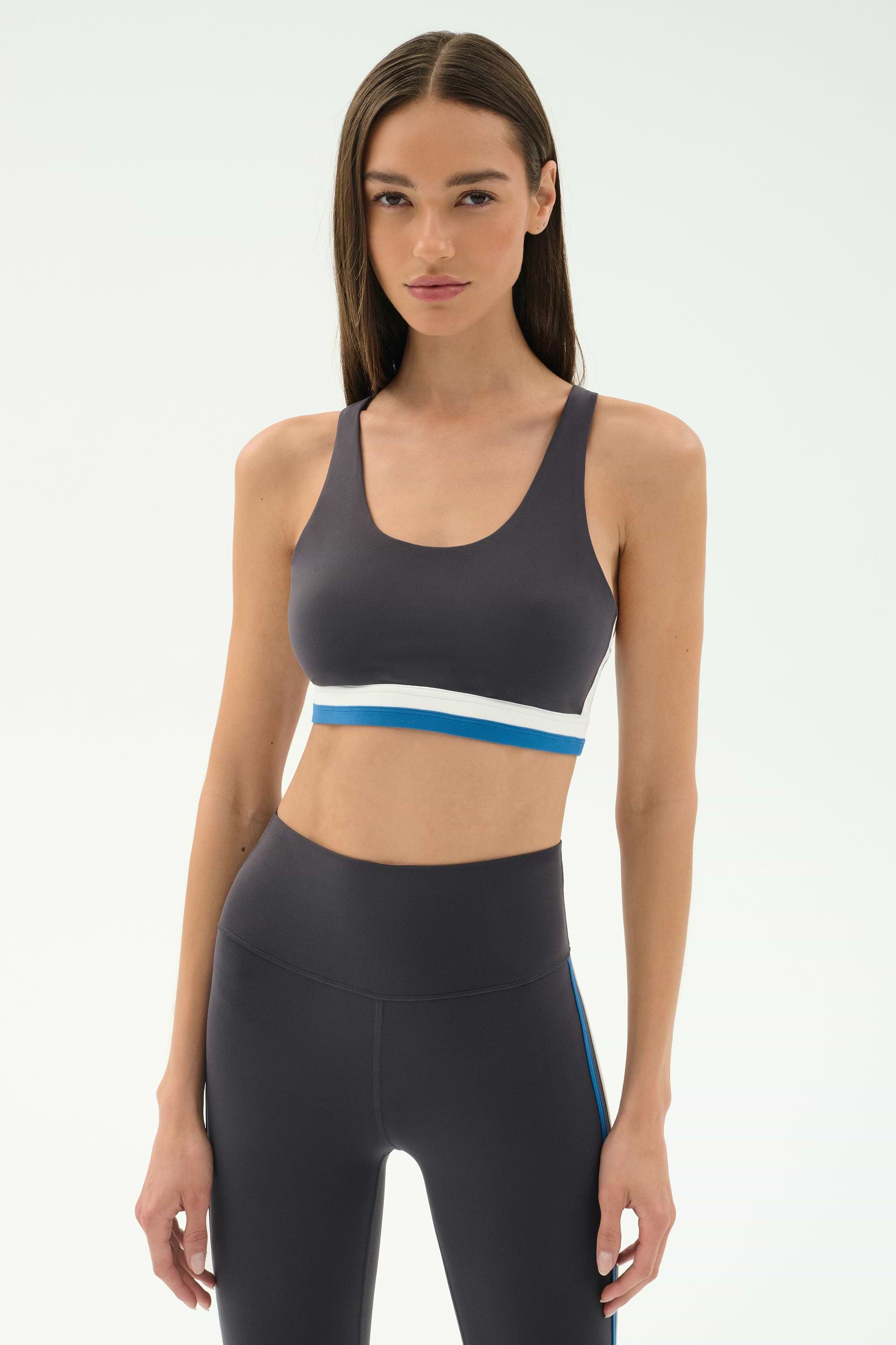 A woman faces forward wearing the SPLITS59 Maci Airweight Bra in Graphite/White with a white and blue band, paired with matching high-waisted leggings, perfect for gym workouts, against a plain light background.