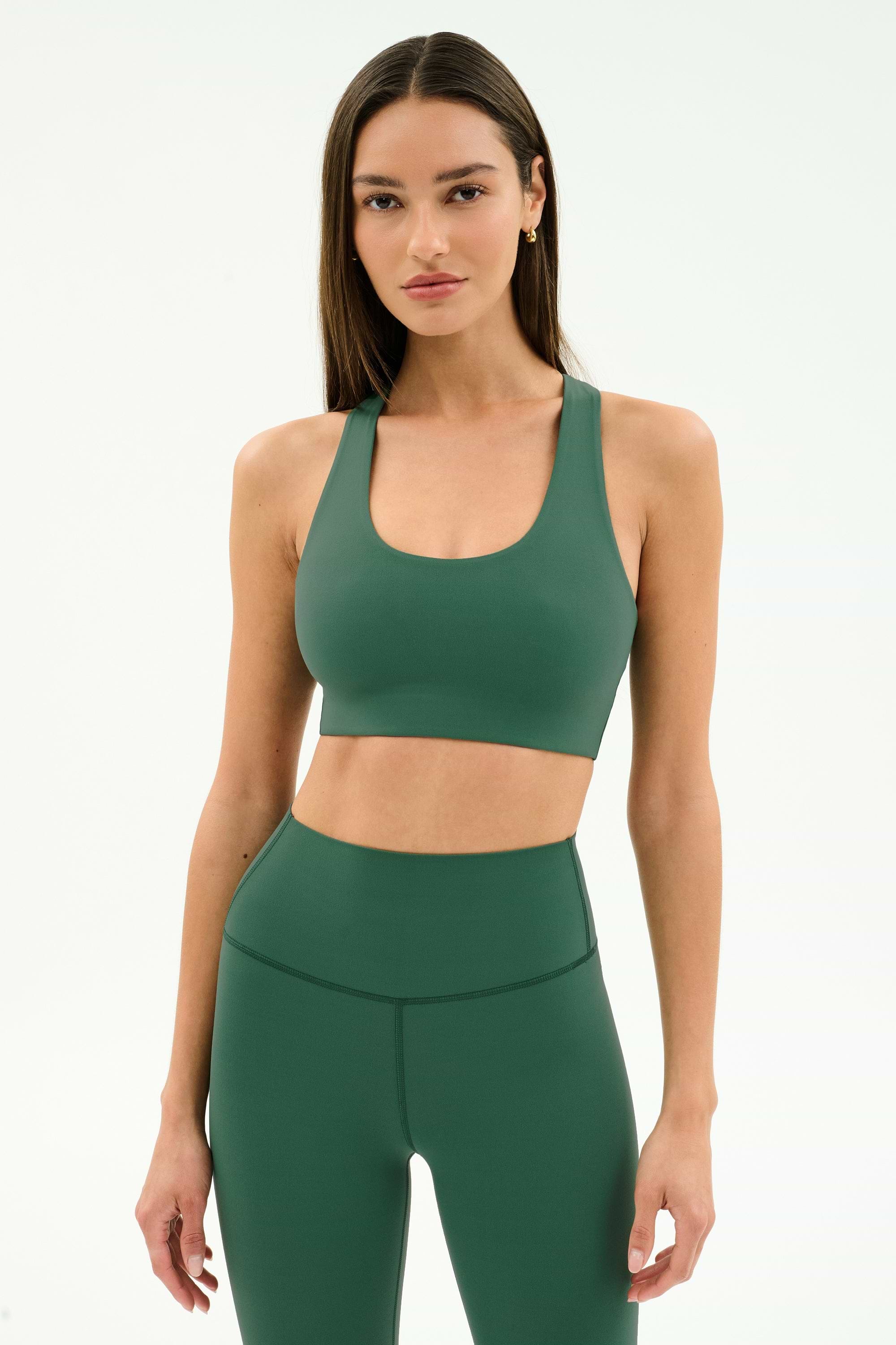 A woman wears the SPLITS59 Sara Airweight Bra in Arugula with matching high-waisted leggings, both crafted from nylon and spandex, standing against a plain light background.