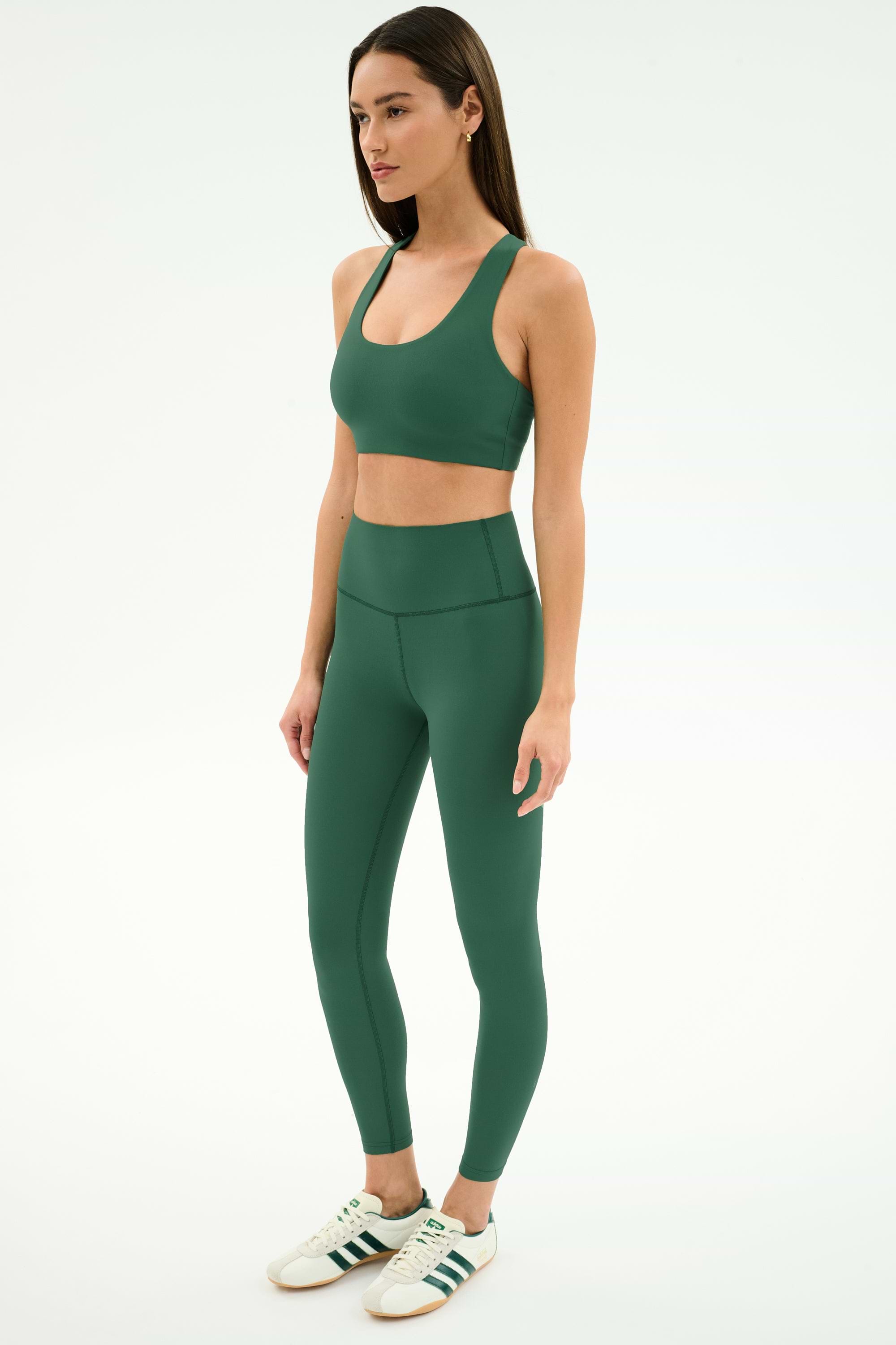 A woman stands against a plain background wearing the SPLITS59 Sara Airweight Bra in Arugula, matching leggings, and white Adidas sneakers with green stripes.