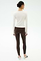 A woman stands with her back to the camera wearing SPLITS59’s Airweight Lite Long Sleeve Waist Length top in white, paired with striped brown leggings and white sneakers—ideal activewear for the gym on a plain light background.