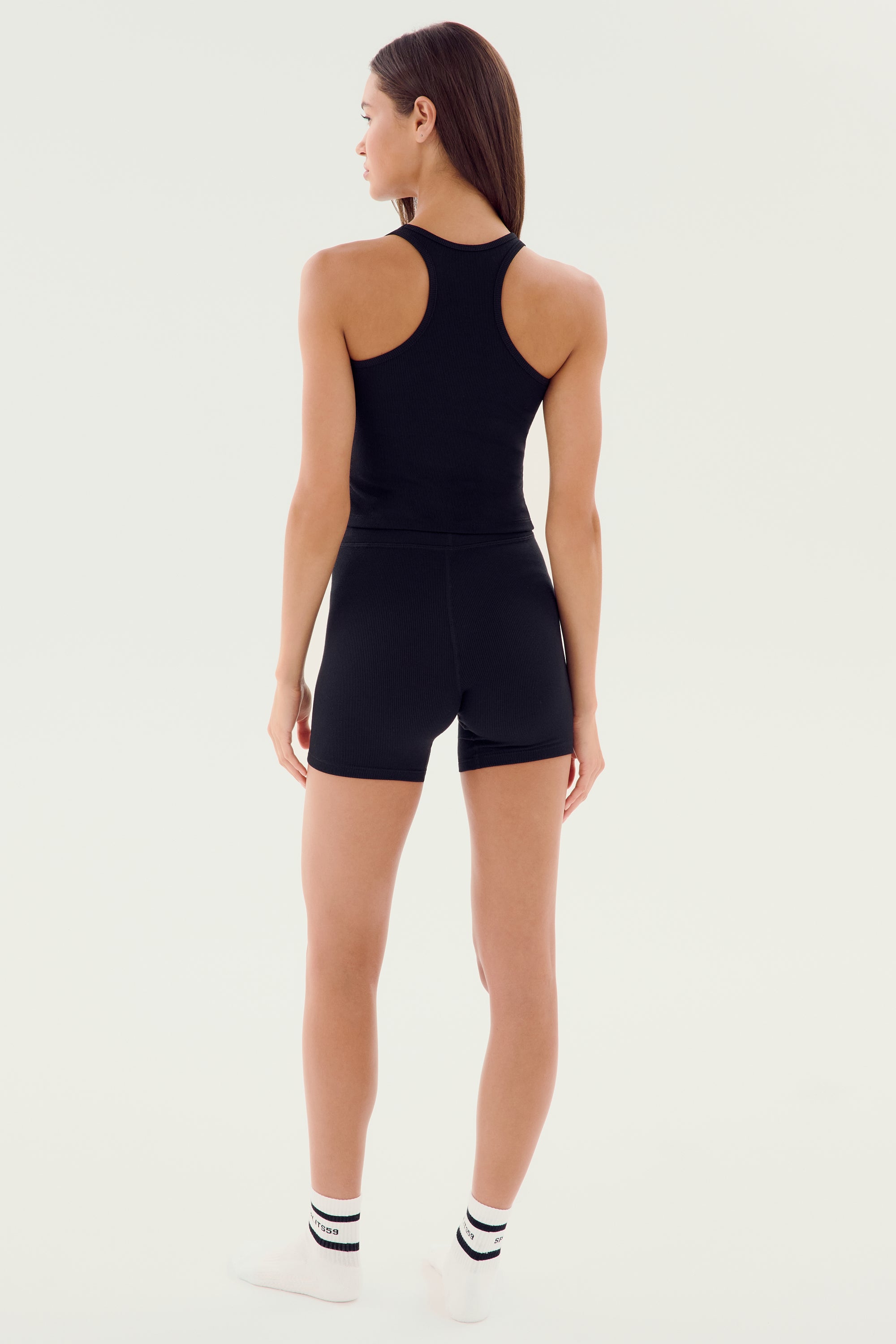 A person with long straight hair stands facing away from the camera, wearing a SPLITS59 Ashby Rib Crop - Black, black shorts, and white ankle socks with black stripes.