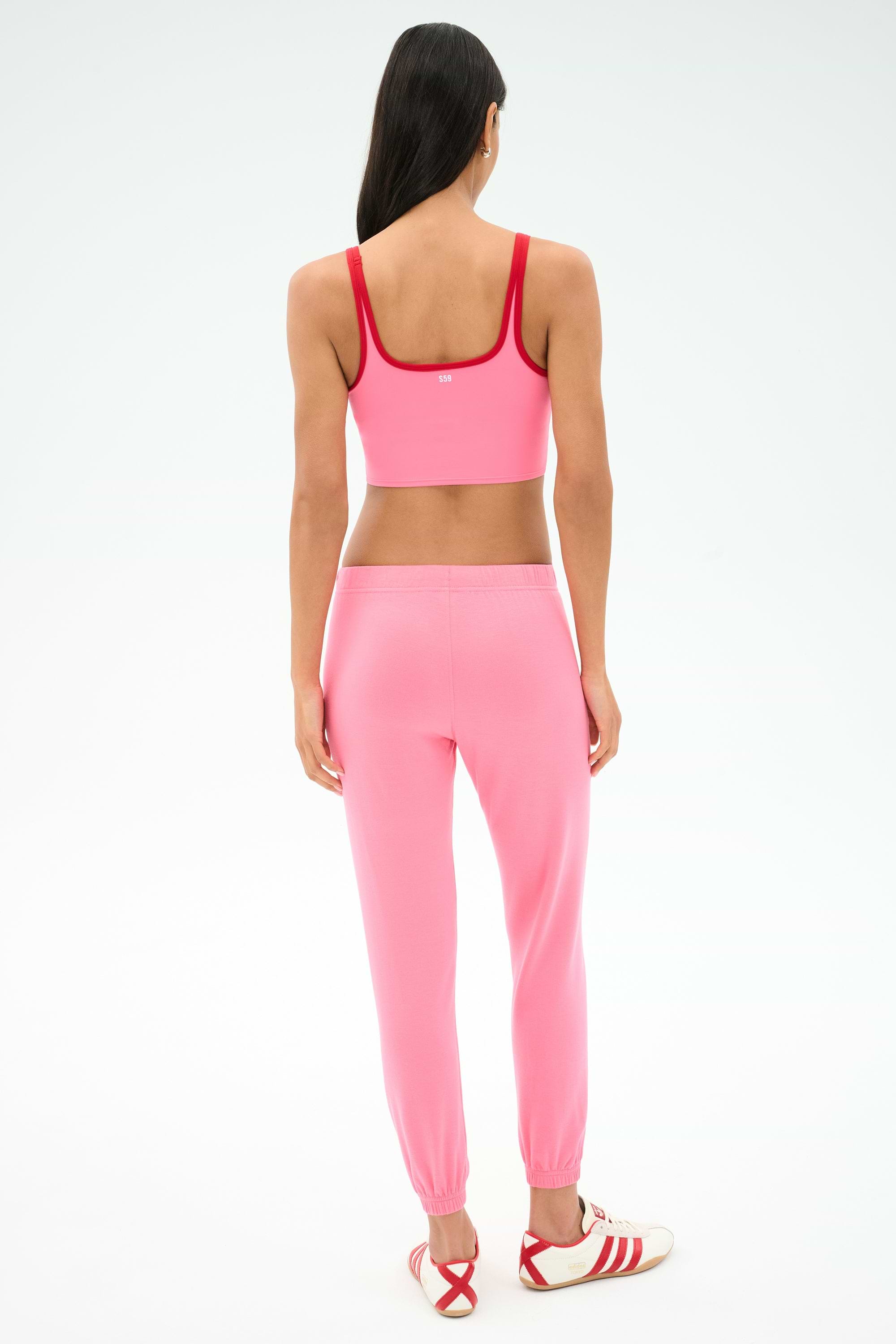 A person stands facing away, wearing the SPLITS59 Sonja Fleece Sweatpant in Strawberry, a pink sports bra with red trim, and white sneakers with red accents.