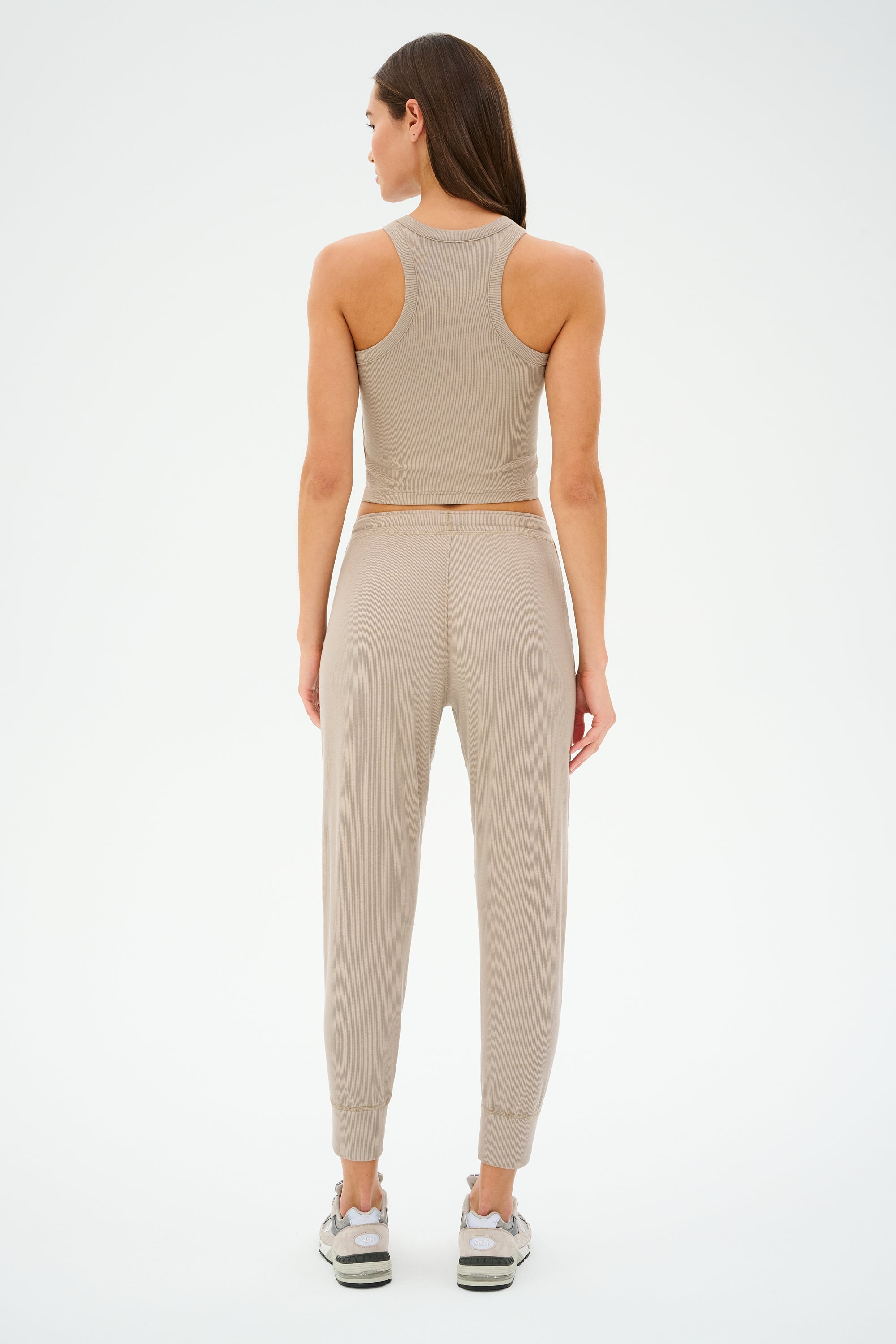 A person stands facing away, wearing the SPLITS59 Kiki Rib Crop Tank - Latte, crafted from baby rib fabric, with matching pants and sneakers against a plain background. This chic MADE IN LOS ANGELES outfit embodies effortless style.