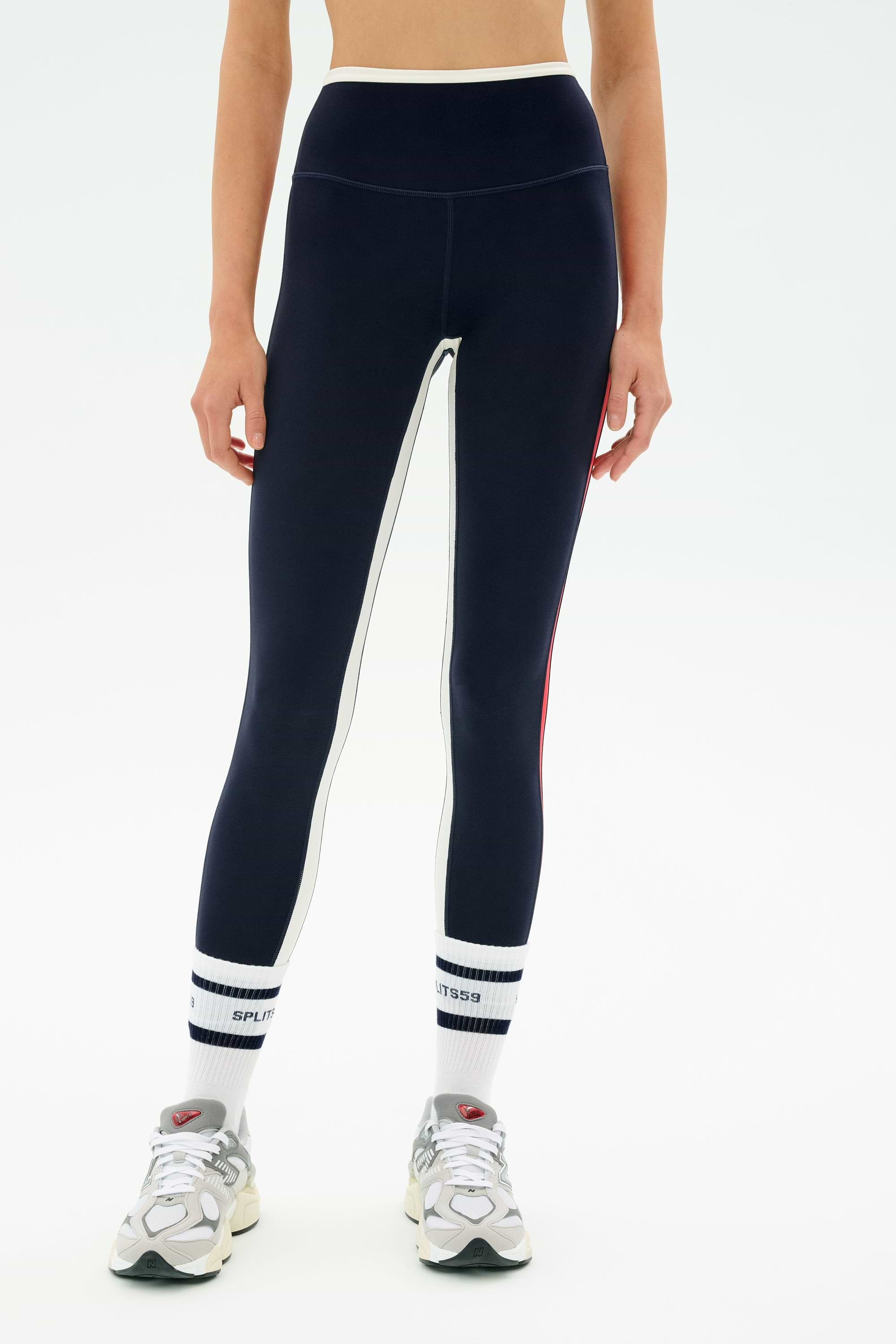 A person wearing SPLITS59 Sam High Waist Rigor 7/8 navy blue leggings with white side stripes, paired with white socks featuring black stripes and gray athletic shoes, stands against a plain white background.