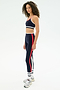 A person stands in a side pose on a plain white background wearing SPLITS59 Sam High Waist Rigor 7/8 leggings in Indigo/White with red and white stripes, paired with a matching sports bra and gray sneakers.