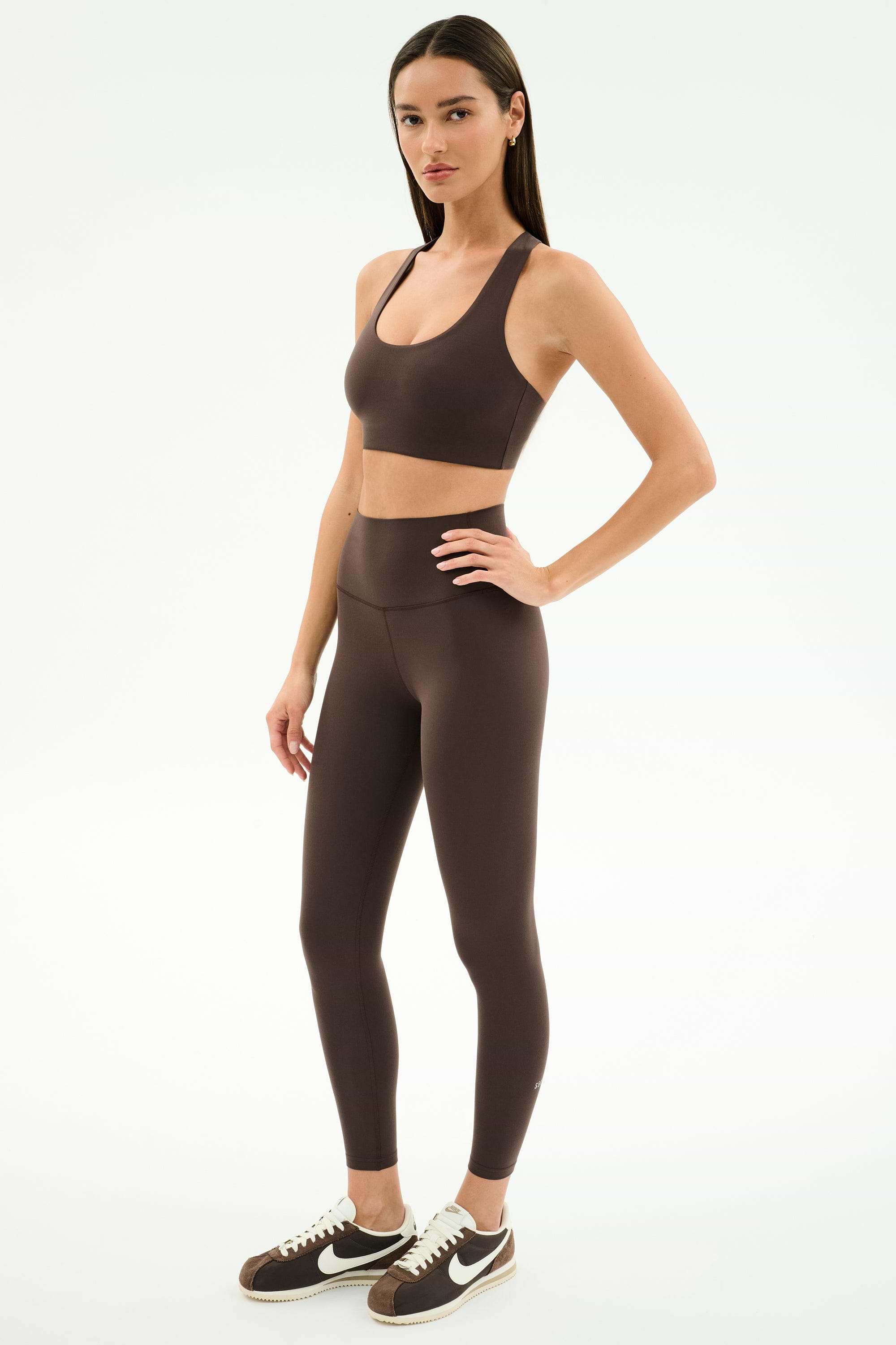 A woman stands in a studio against a white background wearing SPLITS59's Airweight High Waist Legging in Dark Chocolate with a matching brown sports bra and brown Nike sneakers—ideal for hot yoga.
