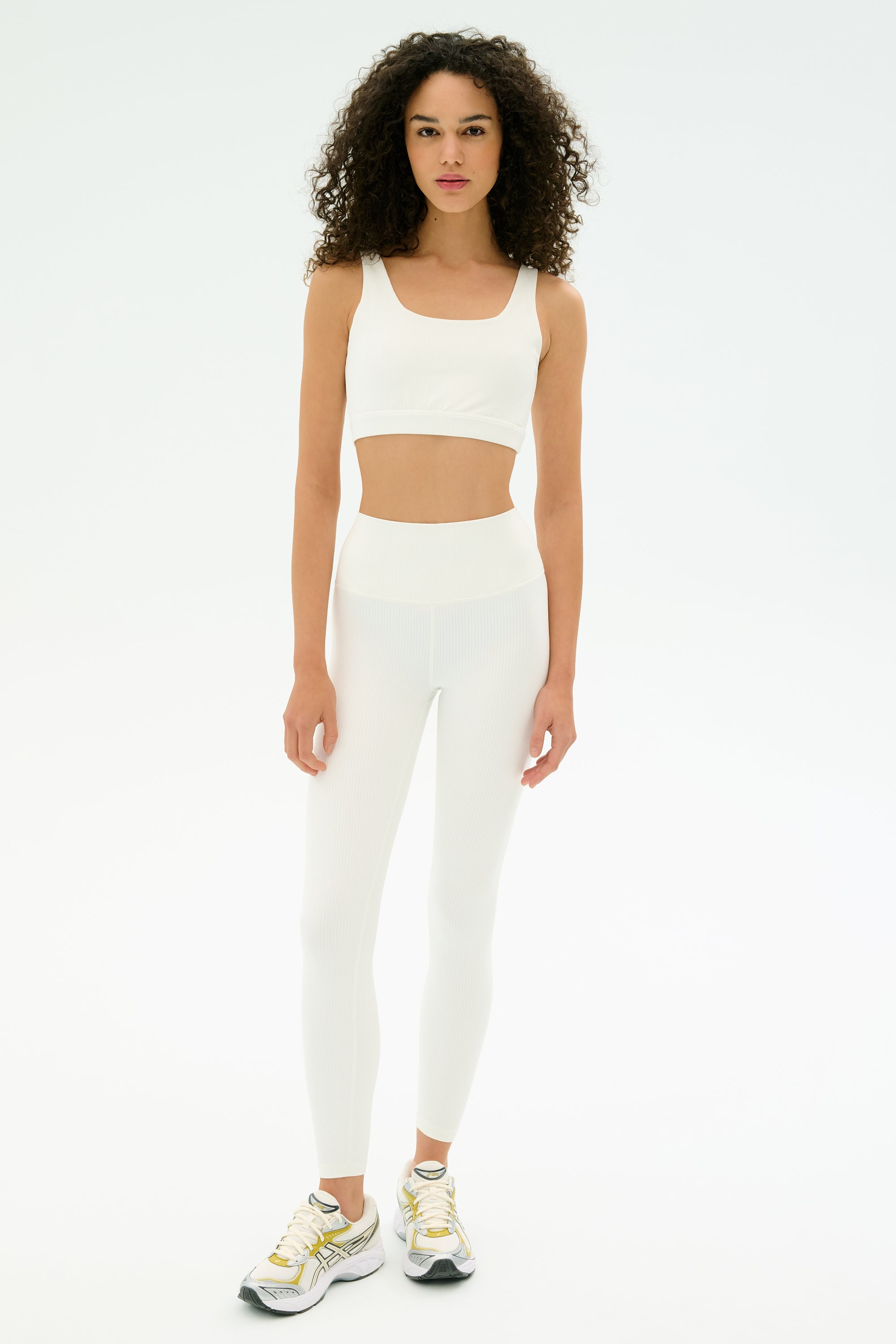 A woman stands on a white background wearing the SPLITS59 Airweight Rib High Waist 7/8 leggings in white and a matching sports bra, paired with silver running shoes.