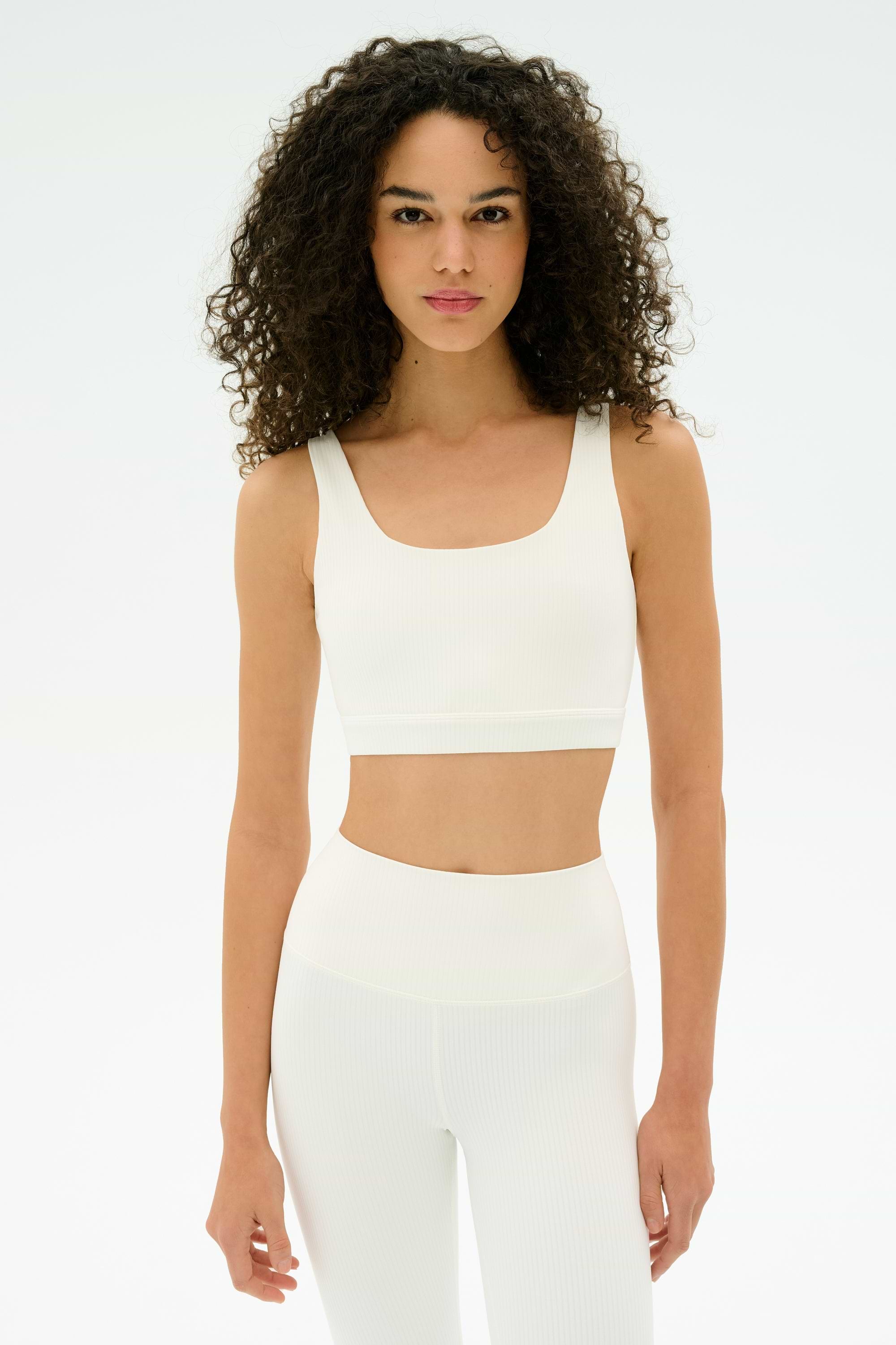 A woman with curly hair wears the SPLITS59 Airweight Rib Bra and matching high-waisted leggings, standing against a plain white background.