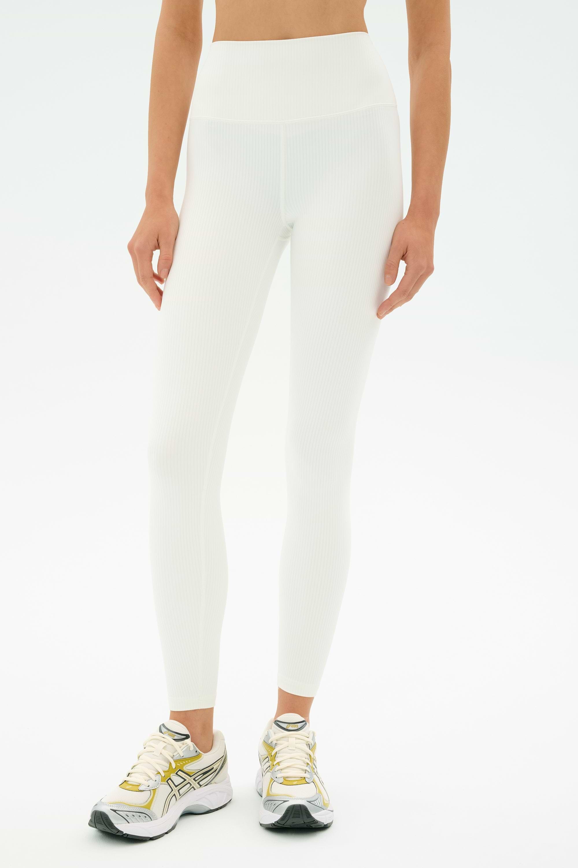 A person wears SPLITS59 Airweight Rib High Waist 7/8 leggings in white and white athletic sneakers with gold and silver accents, standing against a plain white background.