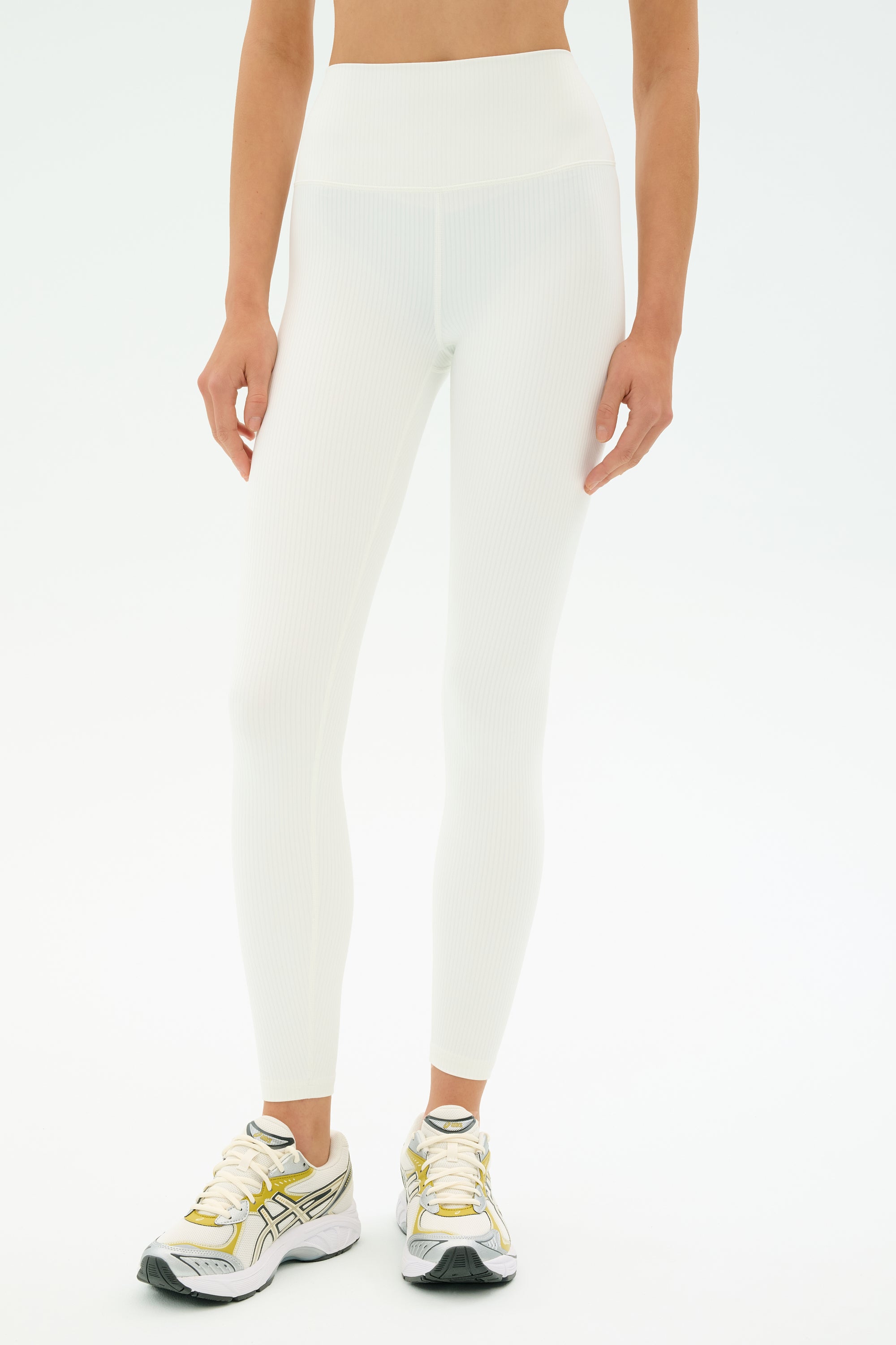 A person wears SPLITS59 Airweight Rib High Waist 7/8 leggings in white and white athletic sneakers with gold and silver accents, standing against a plain white background.