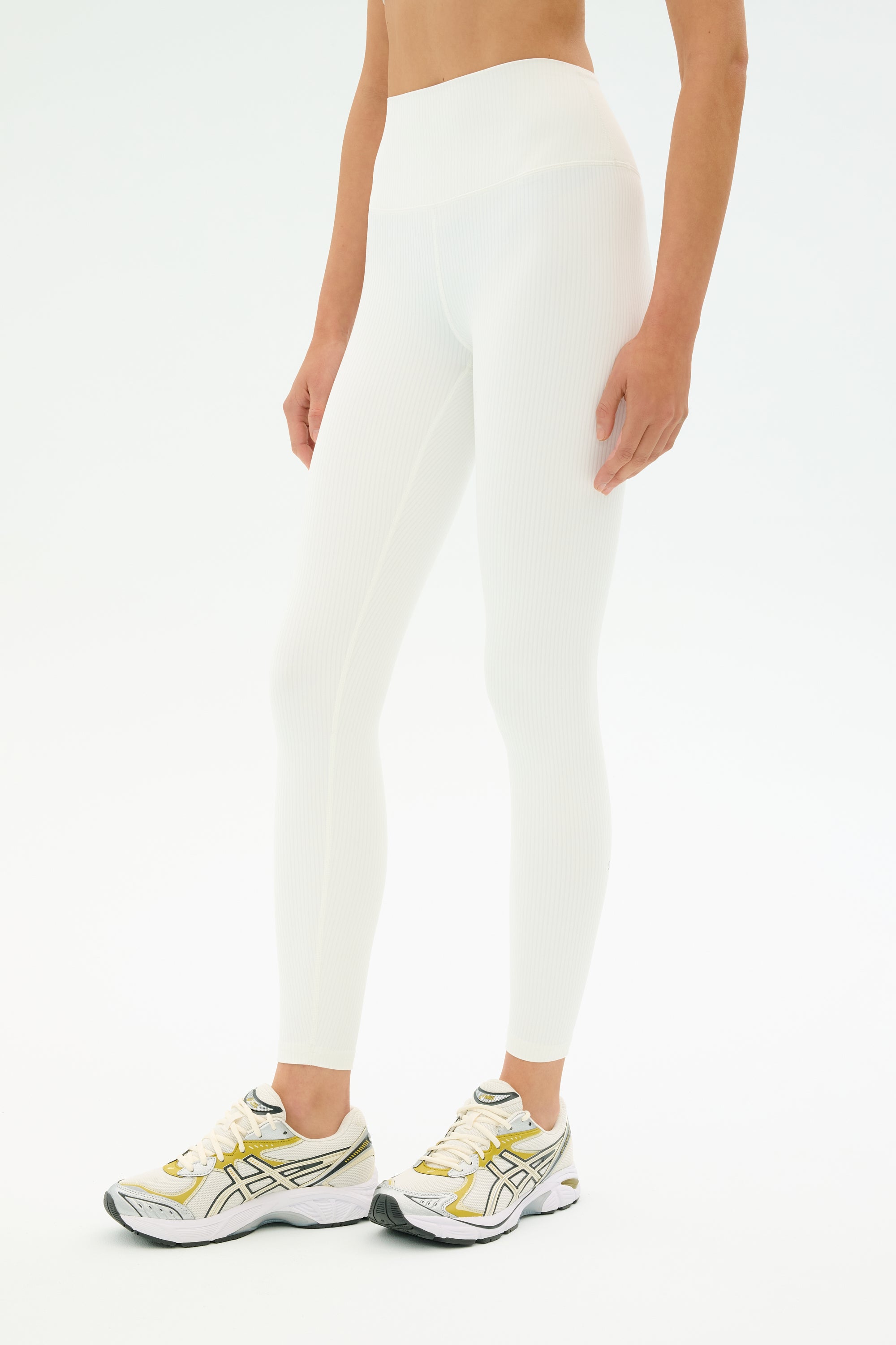 A person wears SPLITS59 Airweight Rib High Waist 7/8 leggings in white with white running shoes featuring yellow and green accents, standing against a plain white background.
