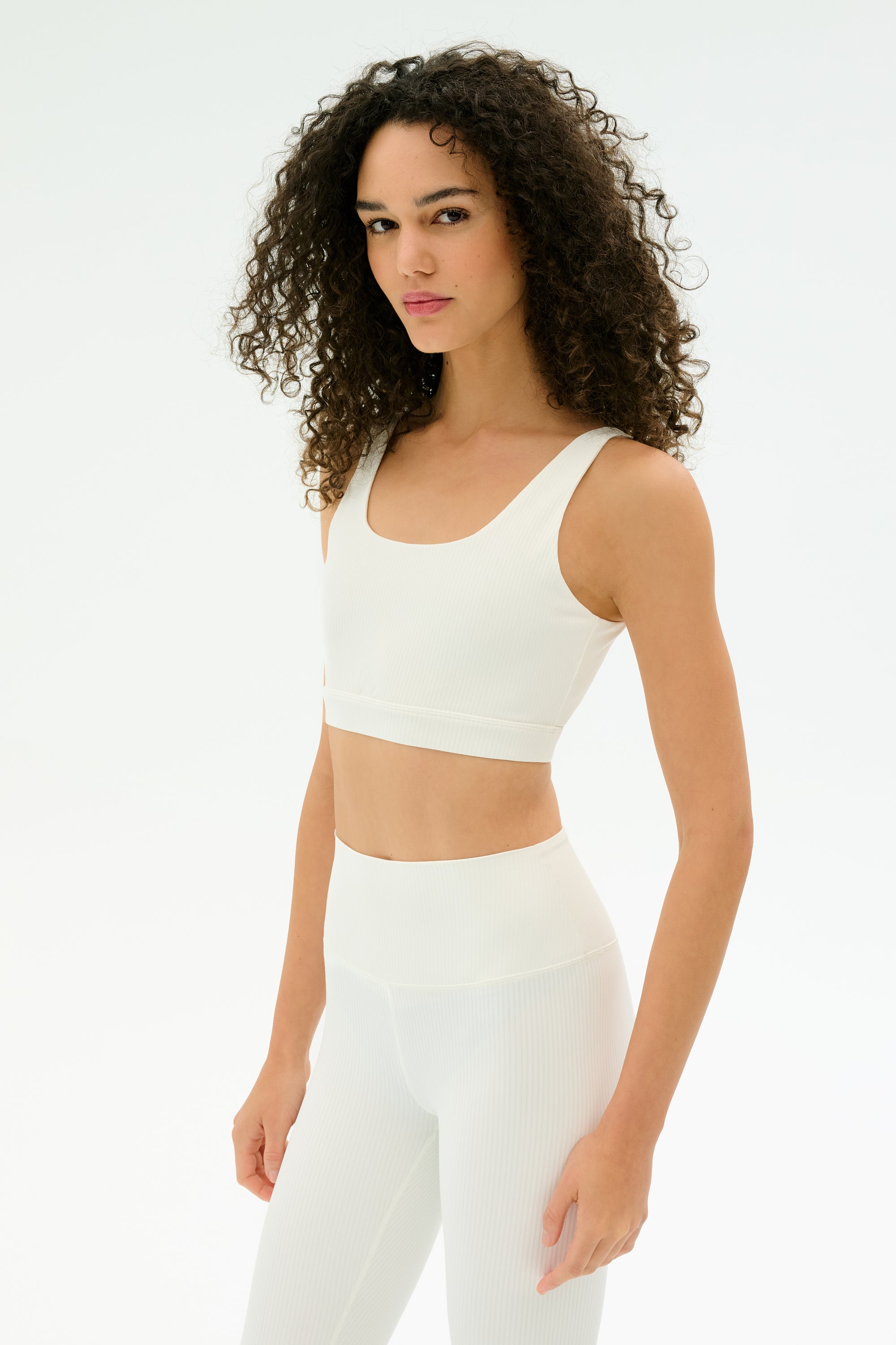A woman with curly hair wears the SPLITS59 Airweight Rib Bra in white, paired with high-waisted leggings, standing against a plain white background.