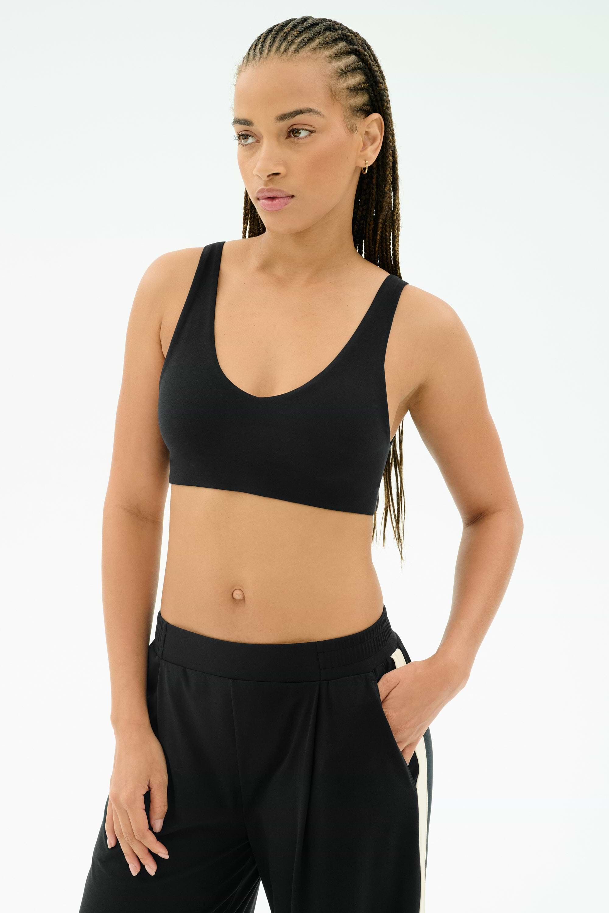 A woman with braided hair wears the SPLITS59 Airweight Scoop Back Bra in black and matching black pants with white stripes, standing against a plain light background.