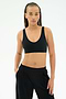 A woman models the SPLITS59 Airweight Scoop Back Bra in black, paired with black pants, standing facing forward with one hand in her pocket against a plain white background.