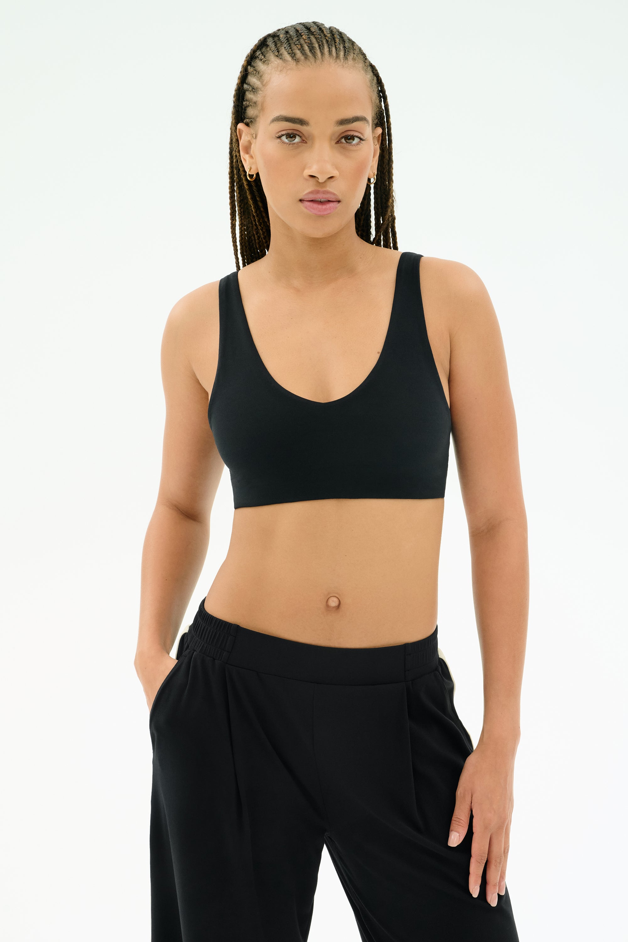 A woman models the SPLITS59 Airweight Scoop Back Bra in black, paired with black pants, standing facing forward with one hand in her pocket against a plain white background.