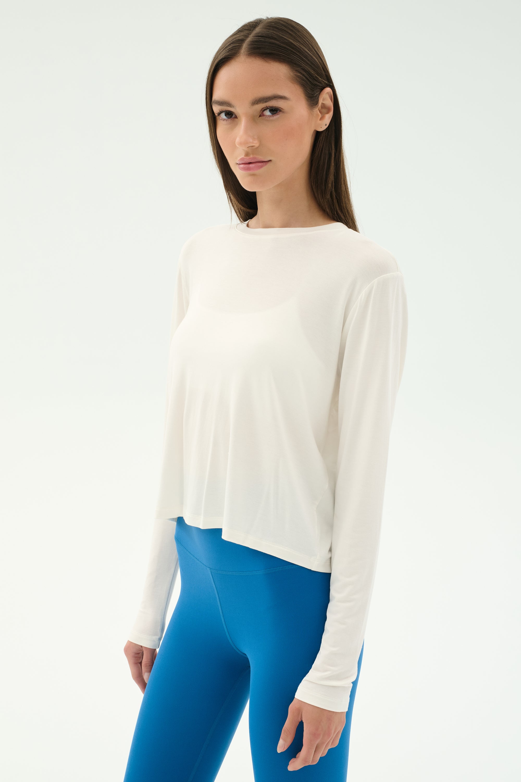 A woman stands against a plain backdrop wearing the SPLITS59 Djuna Long Sleeve in White and bright blue leggings, looking at the camera with a neutral expression—ready for gym or outdoor workouts.