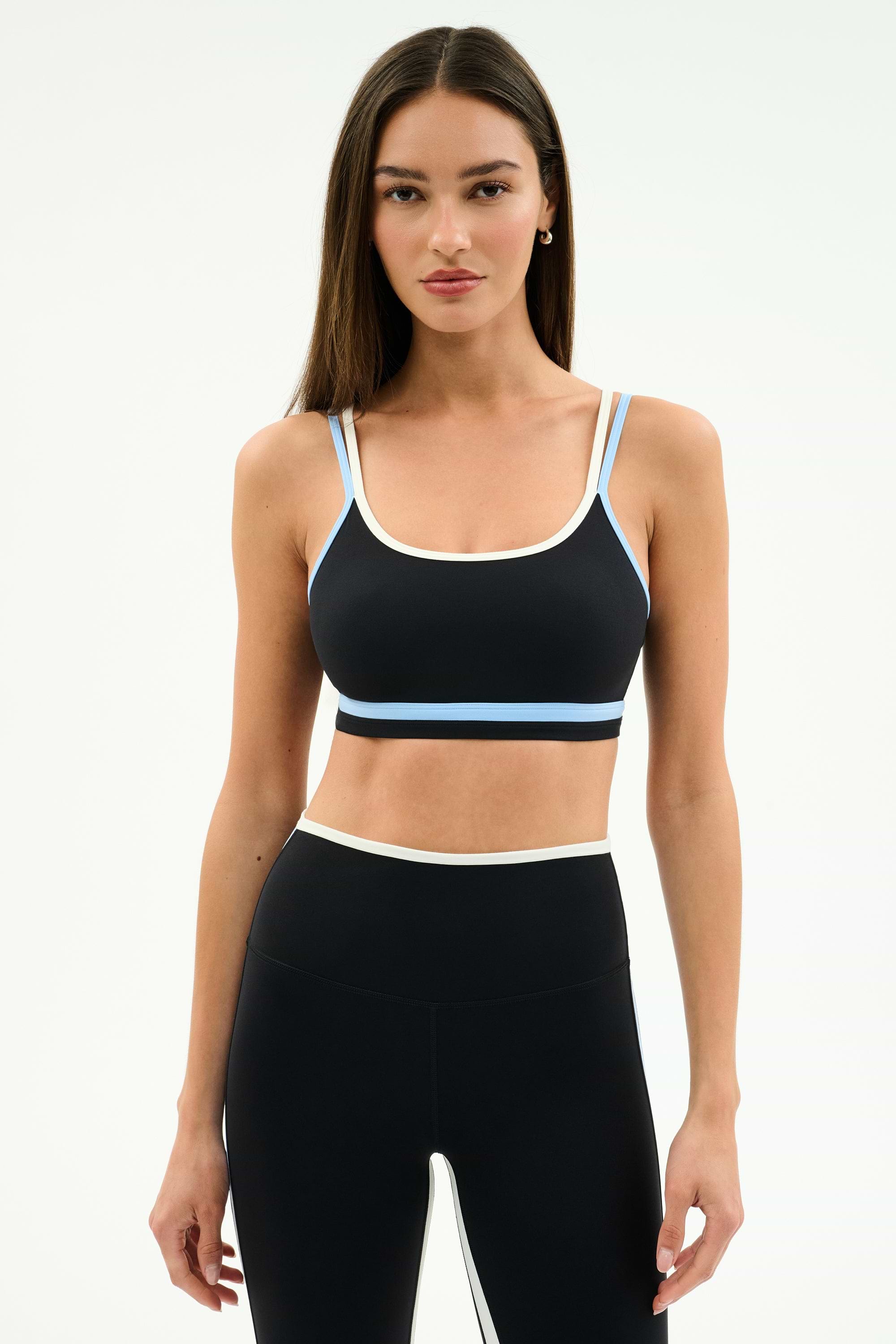 A woman stands against a plain background wearing the SPLITS59 Eva Rigor Double Strap Bra in Black/Skylight with matching high-waisted leggings.