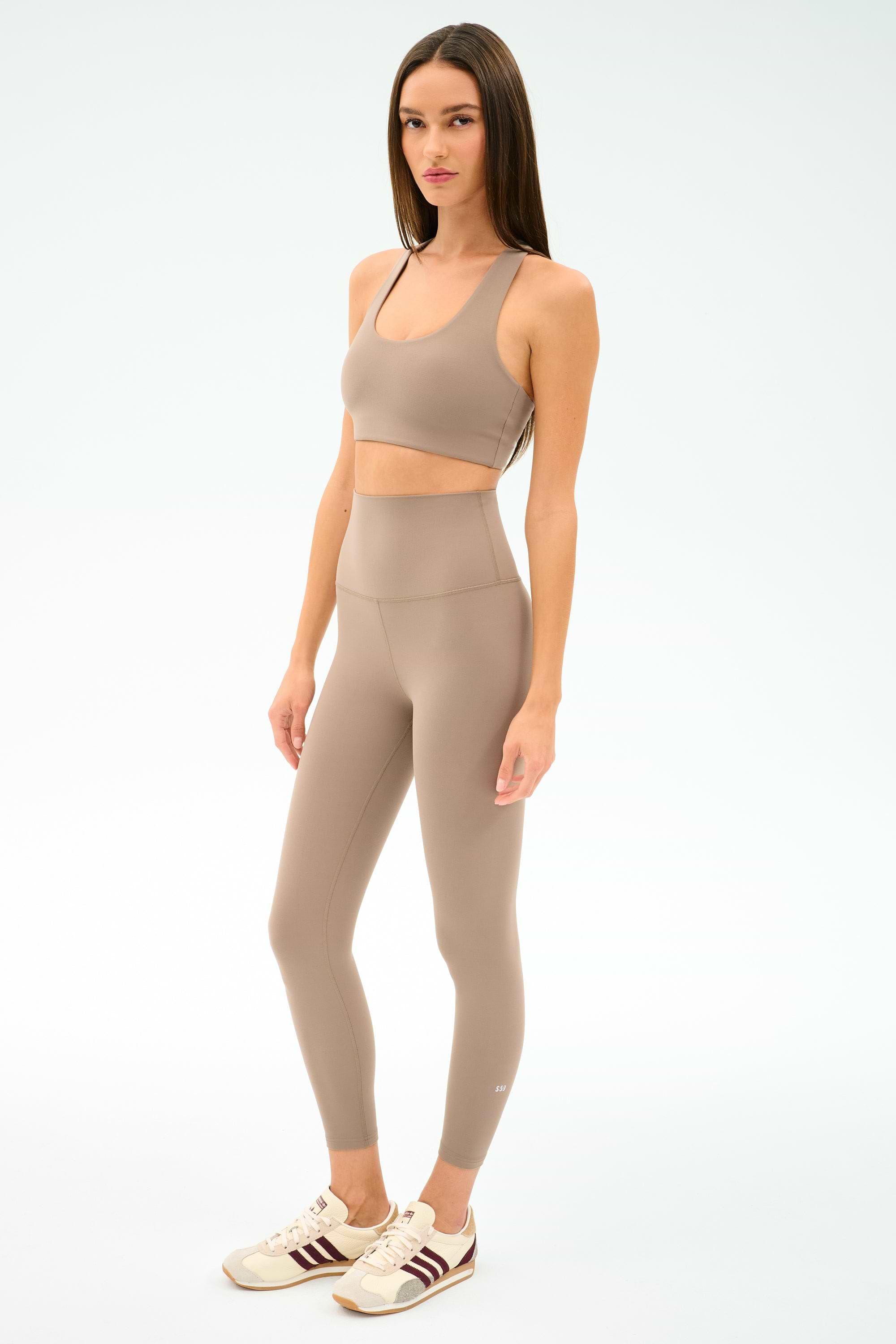 A woman in a SPLITS59 Sara Airweight Bra in Latte stands against a plain white background, ready for Pilates or hot yoga.