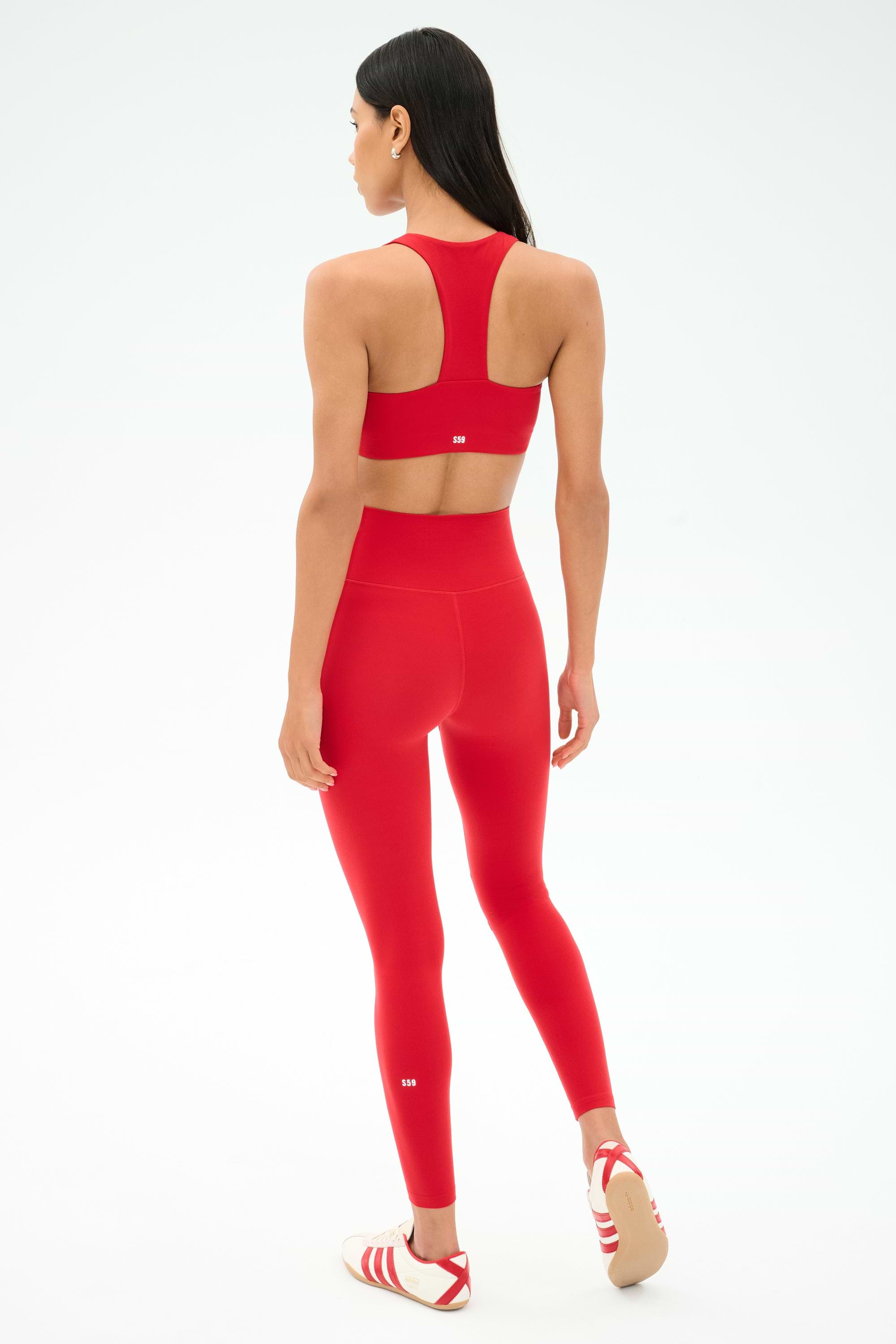 A woman faces away on a white background, wearing SPLITS59’s Airweight High Waist Legging in Pirate Red with a matching red sports bra—ideal for hot yoga—and white/red athletic shoes.