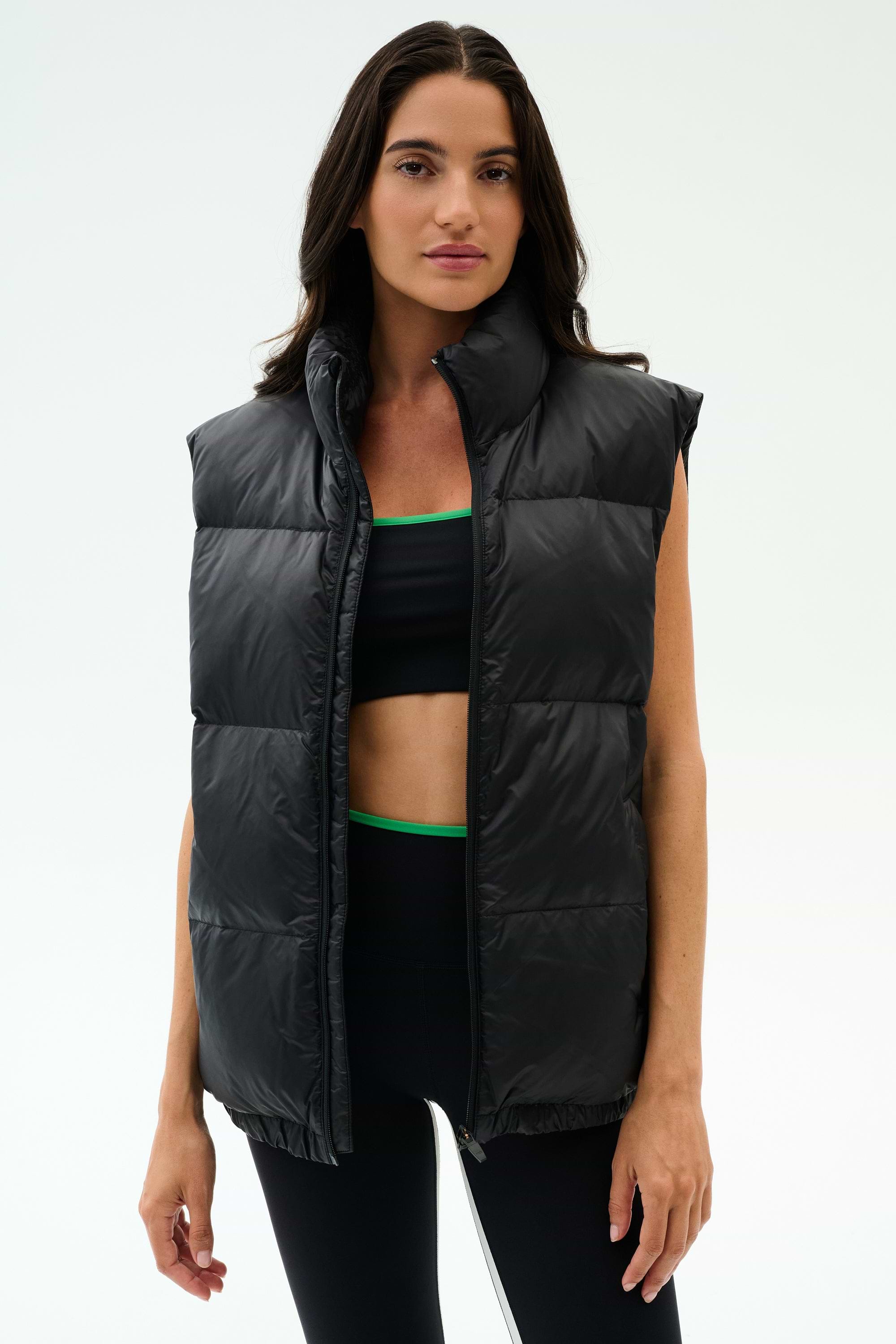 A woman wears the SPLITS59 Summit Nylon Puffer Vest in black over a sports bra and leggings, standing against a plain light background—the perfect lightweight layer for cool weather workouts.