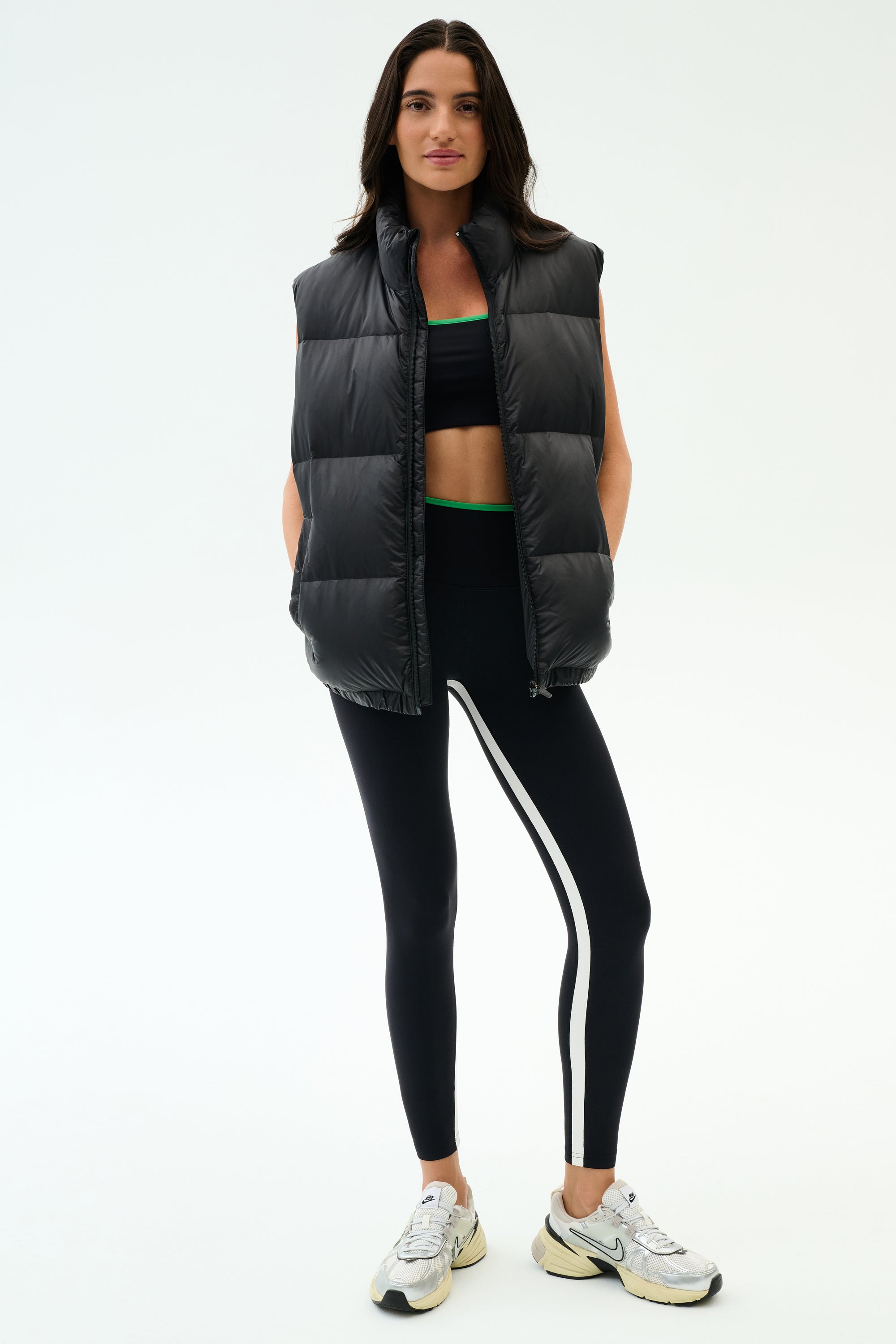 A woman faces forward wearing the SPLITS59 Summit Nylon Puffer Vest in black over a black sports bra, black leggings with a white stripe, and white sneakers—ideal for cool weather workouts against a plain background.