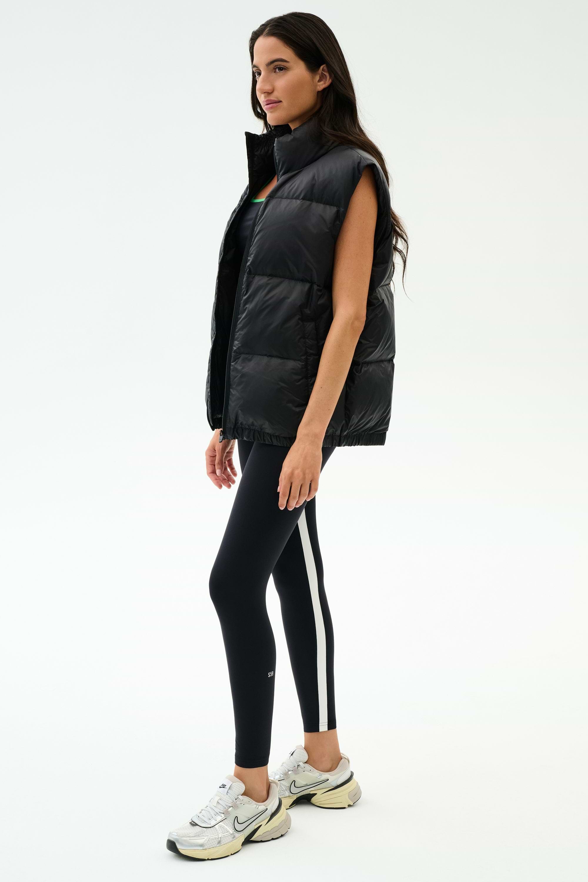 A woman stands in profile against a white background wearing the SPLITS59 Summit Nylon Puffer Vest in black, paired with black leggings featuring a white stripe and white athletic sneakers—highlighting its lightweight, cool-weather insulation for workouts.