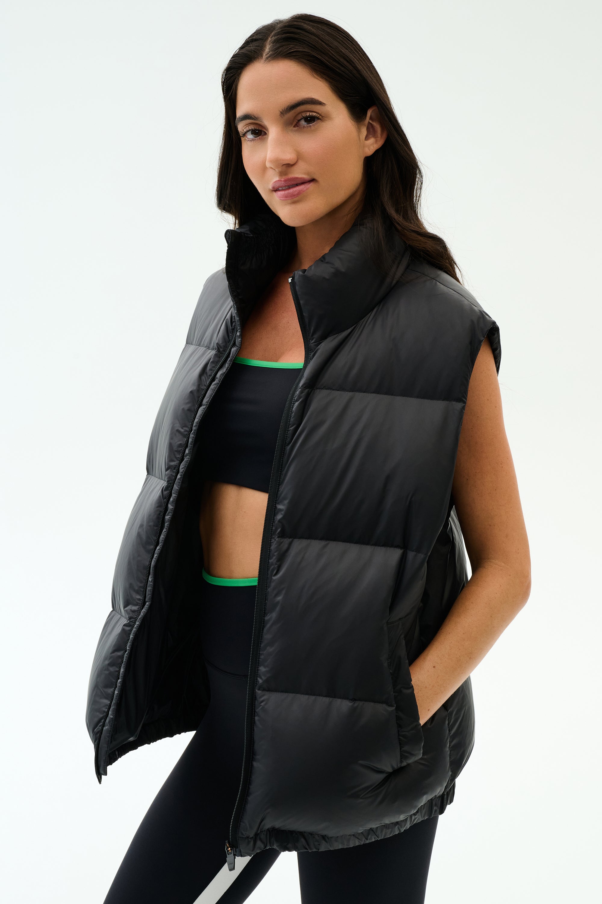 A woman stands against a plain light background wearing the SPLITS59 Summit Nylon Puffer Vest in black over a sports bra and leggings, ready for cool weather workouts with its lightweight insulation.