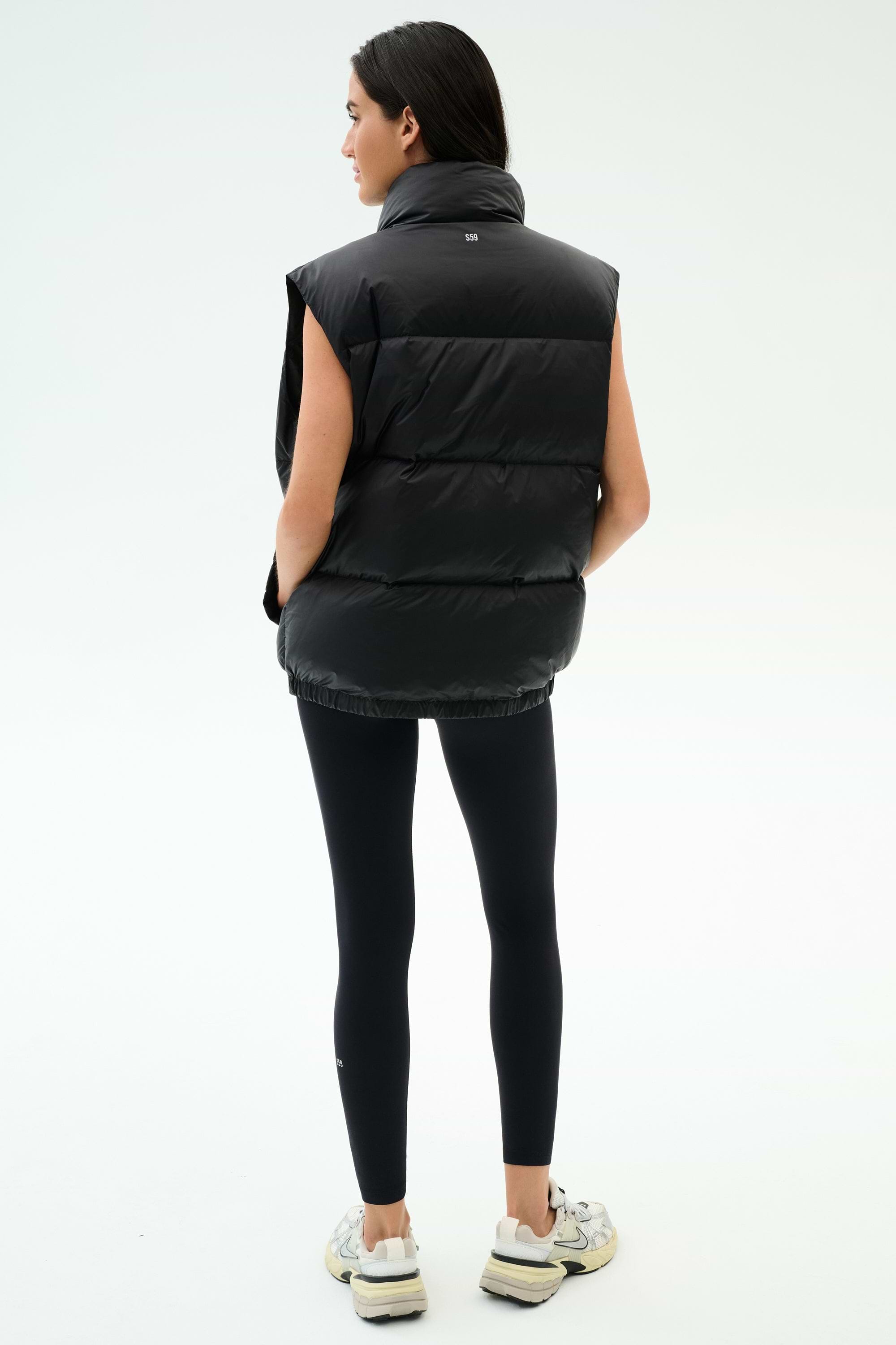 A woman faces away, wearing the SPLITS59 Summit Nylon Puffer Vest in black with lightweight insulation, paired with black leggings and white sneakers against a plain light backdrop—perfect for cool weather workouts.