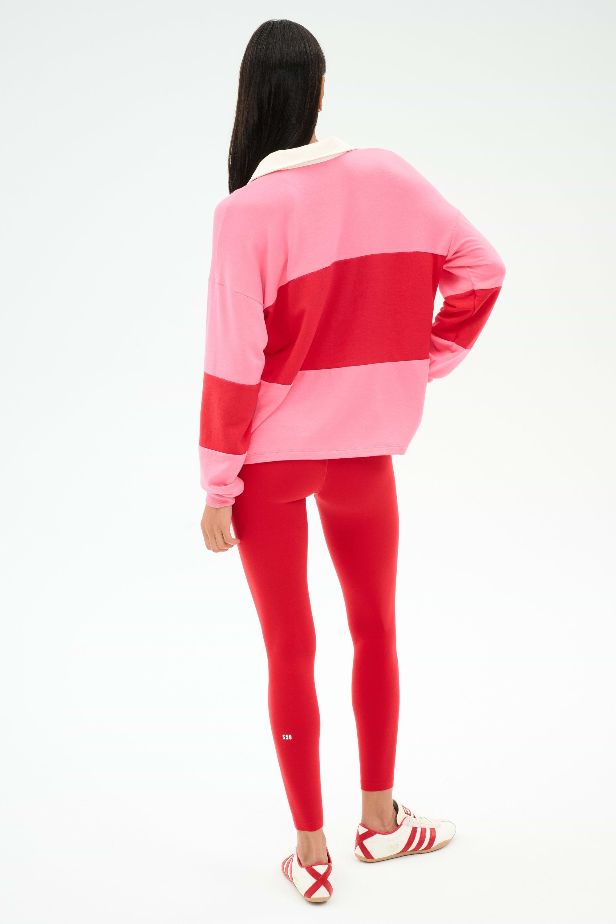 A person with long dark hair faces away, wearing the SPLITS59 Katie Fleece Rugby in Strawberry/Pirate Red, red leggings, and white sneakers with red stripes.