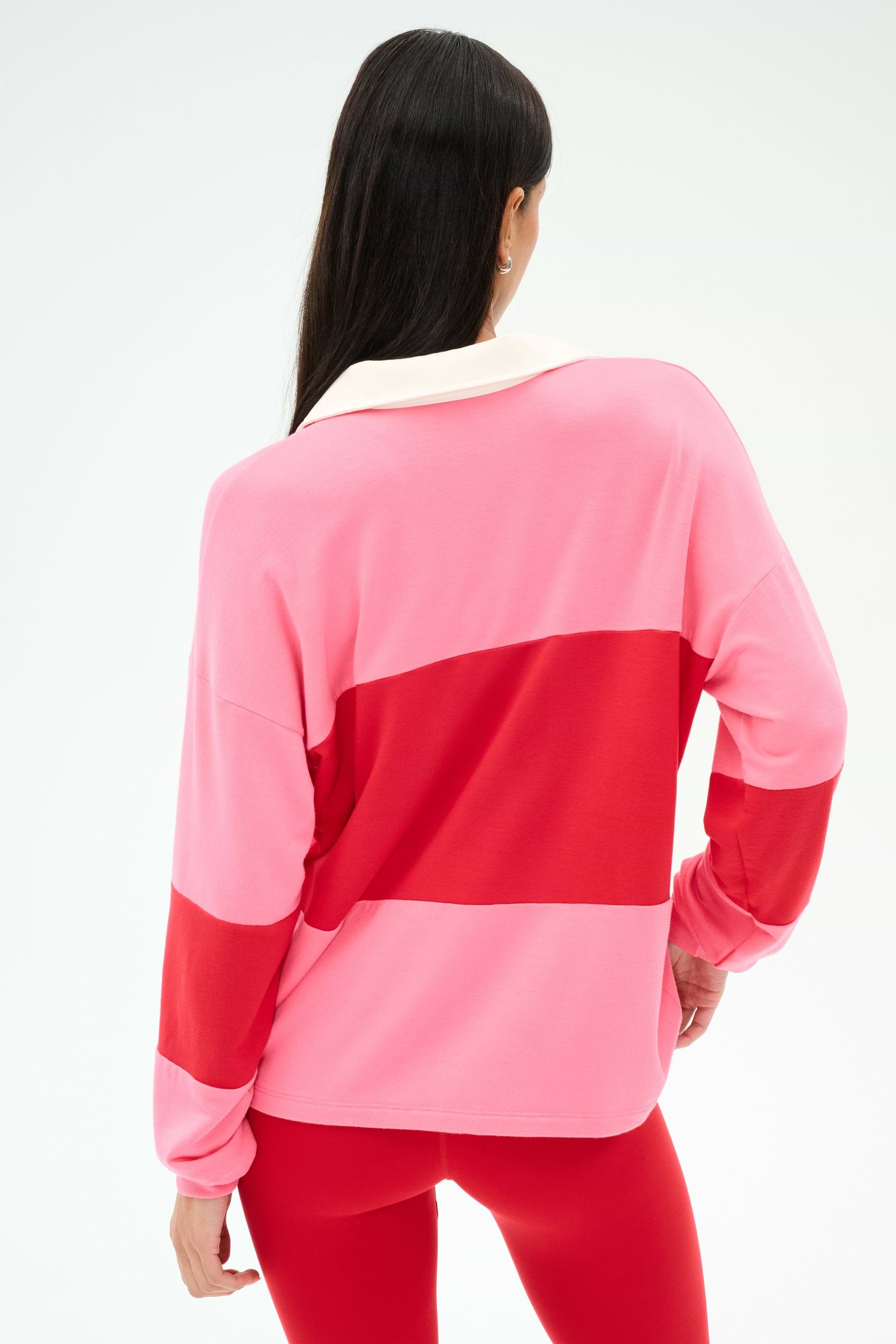 A woman faces away, wearing the SPLITS59 Katie Fleece Rugby in Strawberry/Pirate Red—long-sleeve with wide pink and red stripes, a white collar—and red pants.