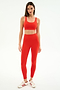 A woman stands against a white background wearing SPLITS59’s Airweight Rib High Waist 7/8 leggings in Lotus Red with a matching sports bra and coordinated red and white sneakers.