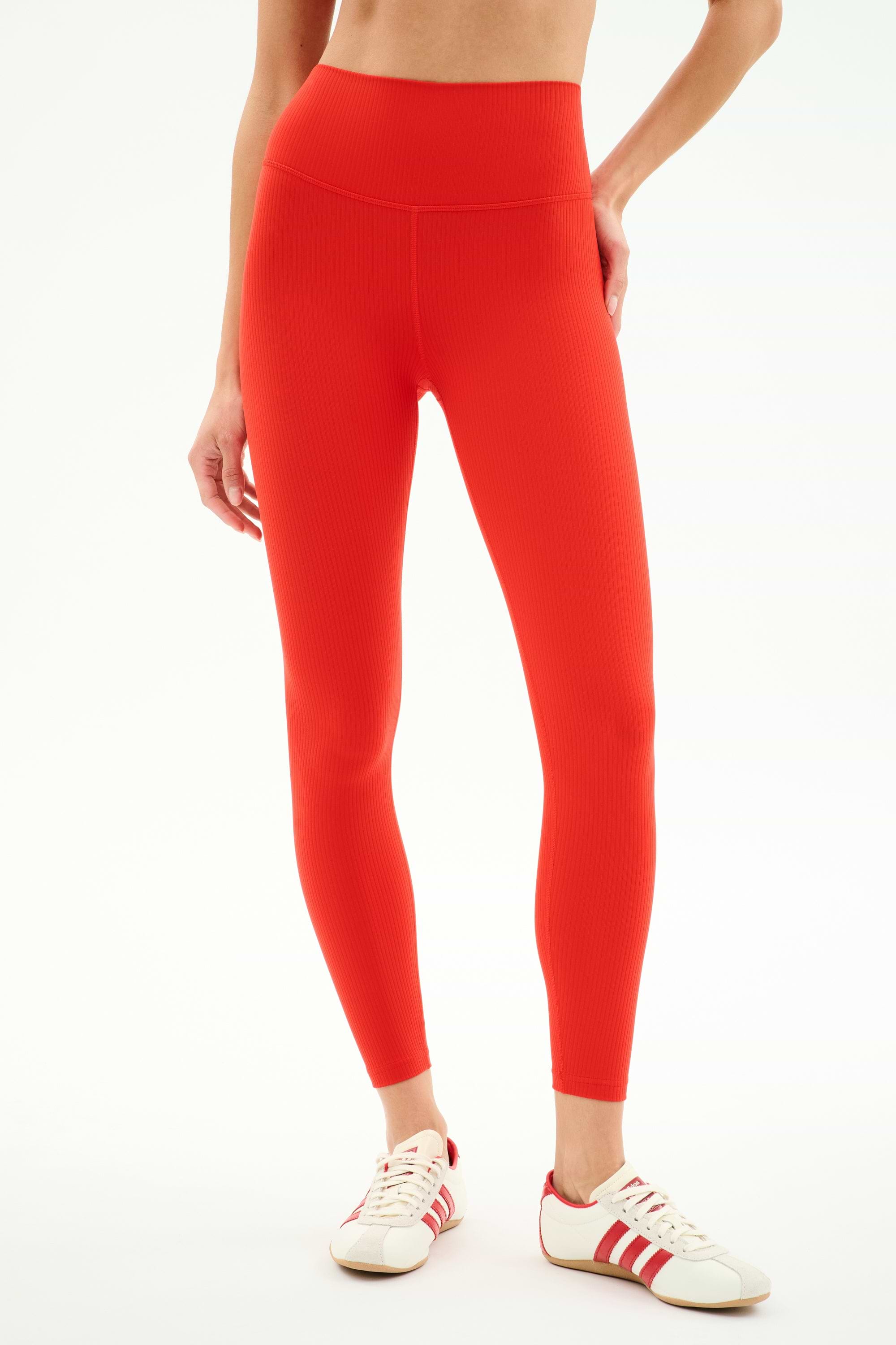 A person wearing SPLITS59 Airweight Rib High Waist 7/8 leggings in Lotus Red with red and white sneakers stands against a plain white background.
