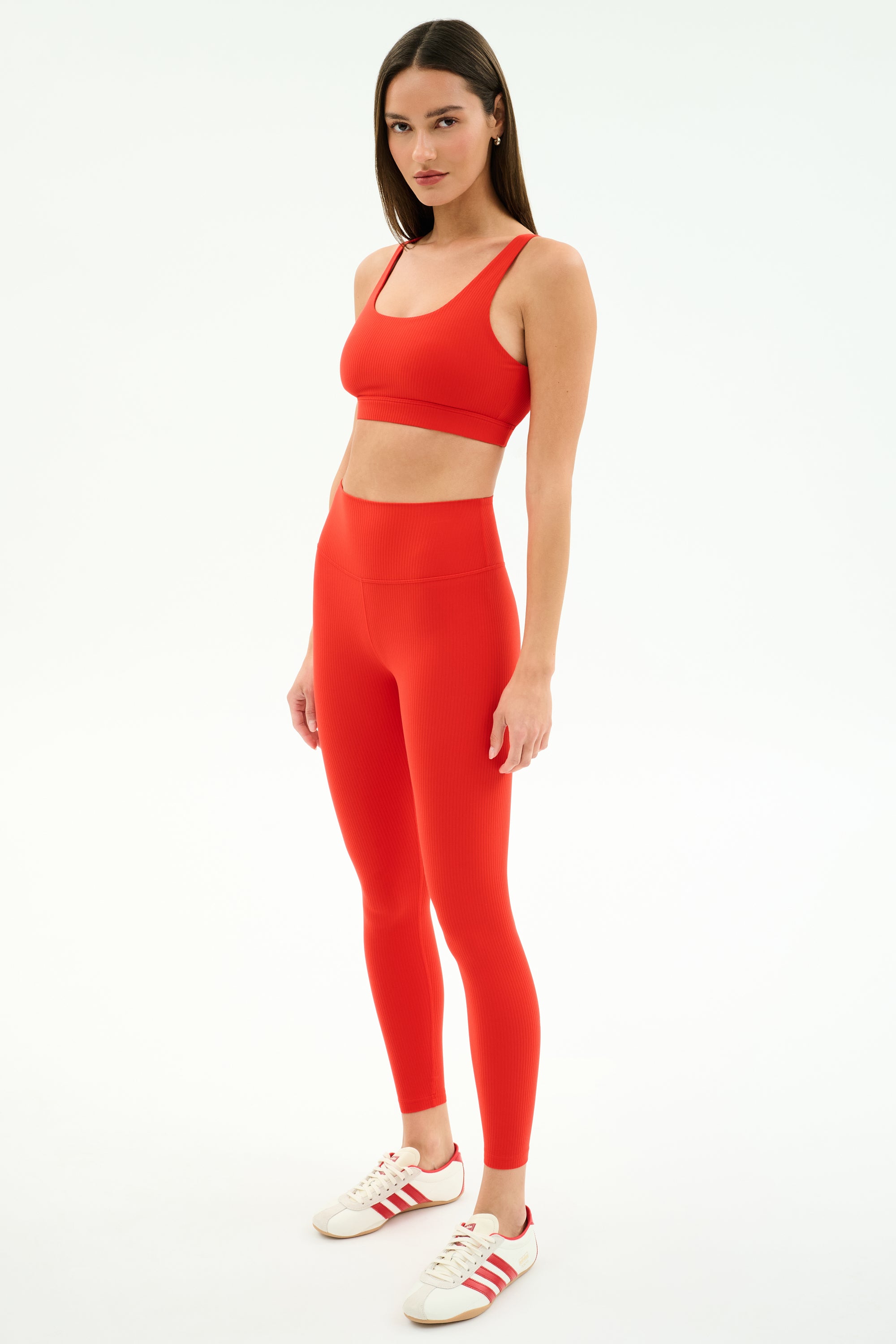 A woman poses against a plain background in the SPLITS59 Airweight Rib Bra - Lotus Red, paired with matching leggings and white sneakers with red stripes—an ideal set for hot yoga.