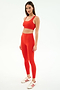 A woman stands against a white background wearing SPLITS59 Airweight Rib High Waist 7/8 leggings in Lotus Red, paired with a matching red sports bra and white-red sneakers.