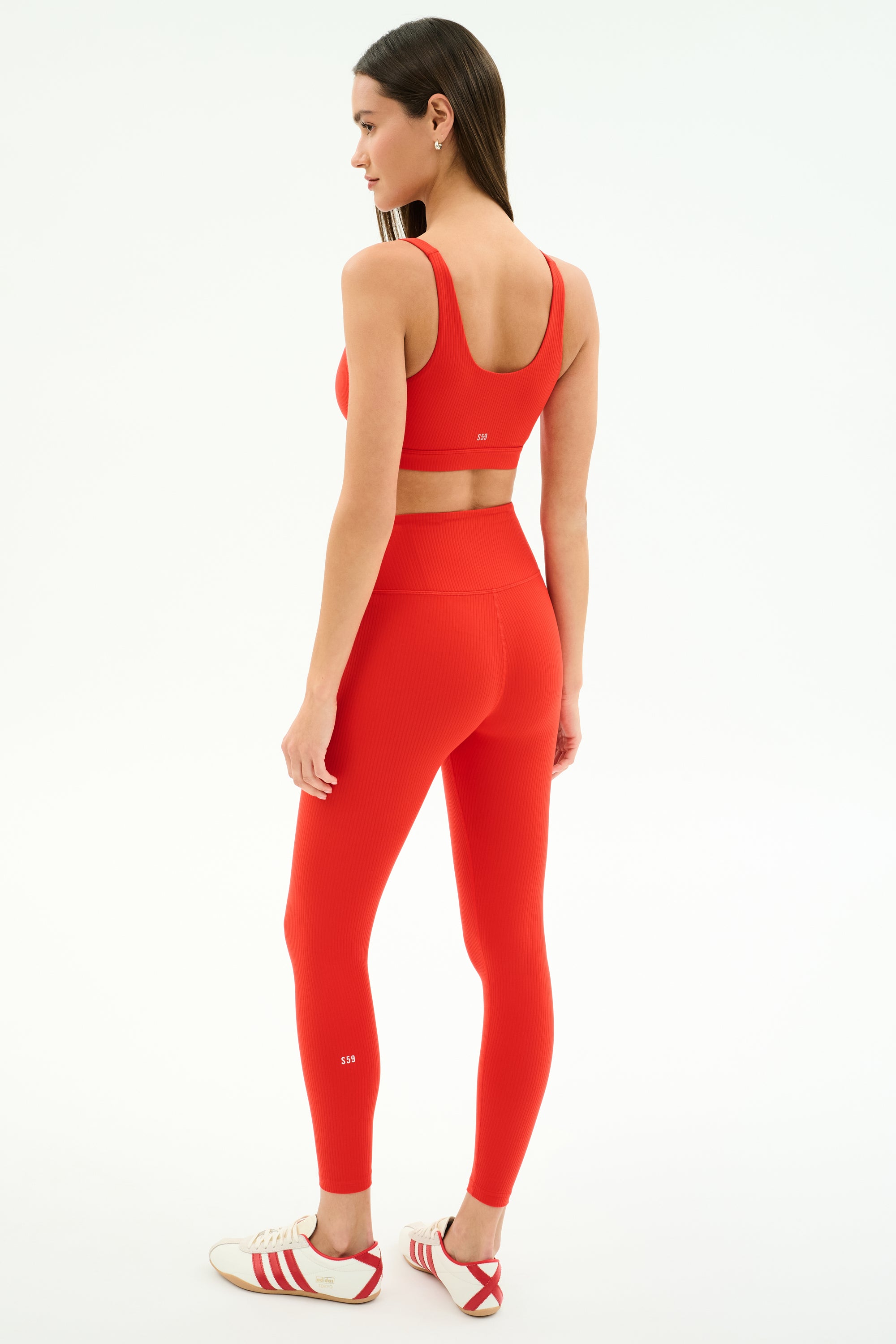 A woman faces away, wearing the SPLITS59 Airweight Rib Bra in Lotus Red with matching leggings and white Adidas sneakers with red stripes, set against a plain white background.